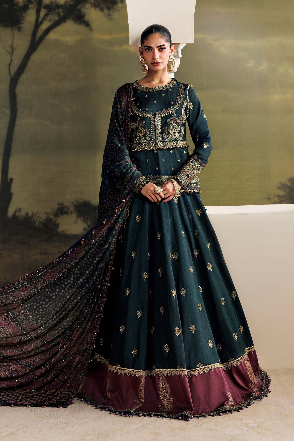 A woman models a dark teal green traditional Indian anarkali gown with gold embroidery, a maroon border, and a matching sheer dupatta, set against a scenic painted backdrop.