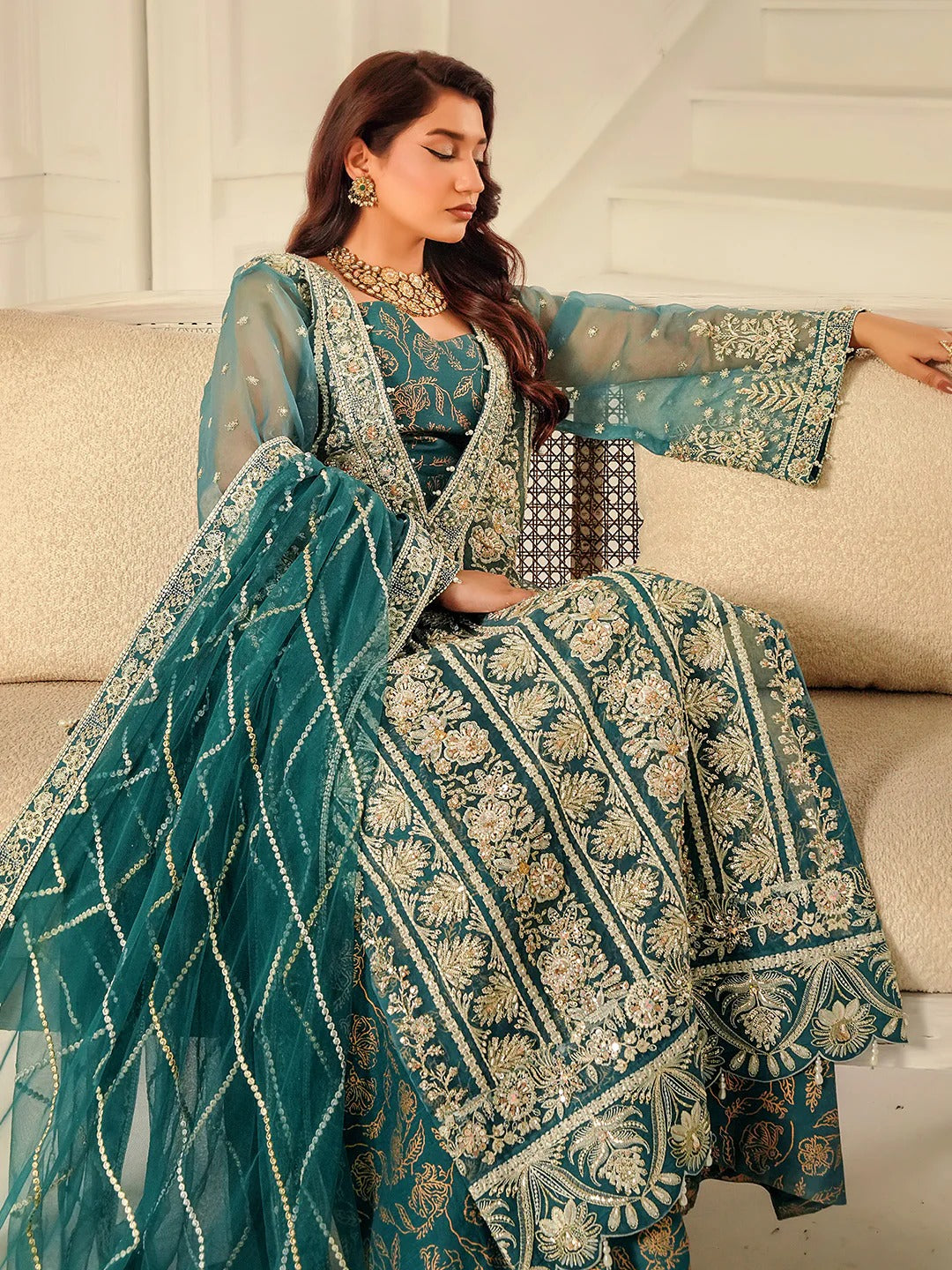 Woman in a teal embroidered traditional outfit sitting on a beige couch.