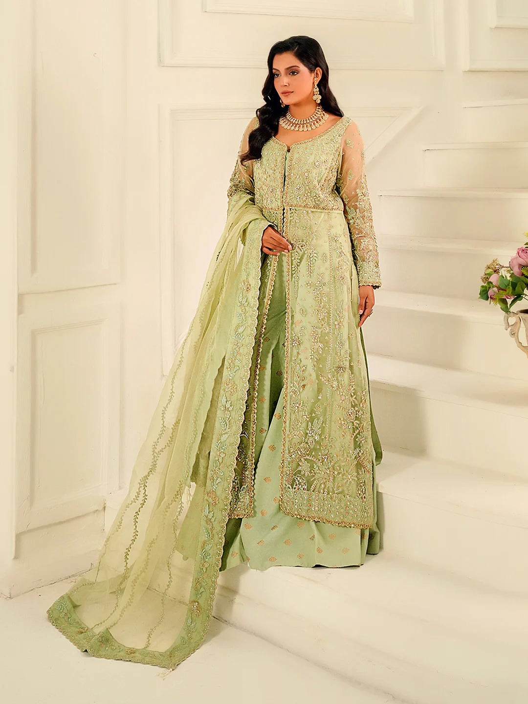 Woman in a light green embroidered traditional outfit with a matching dupatta in a white interior setting.