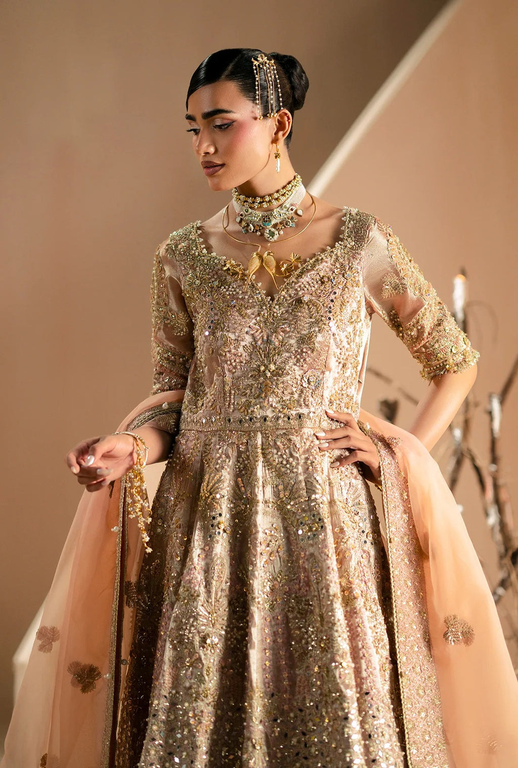 A woman models an ornate traditional Anarkali gown in shimmering gold and beige with intricate sequin embroidery, paired with a sheer peach dupatta and heavy gold jewelry against a warm beige background.