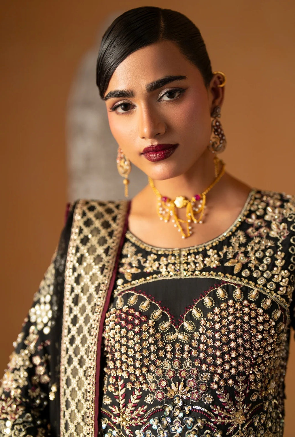 A close-up portrait of a woman wearing an ornate black and gold embellished traditional outfit with heavy beadwork, paired with gold jewelry against a warm brown background.