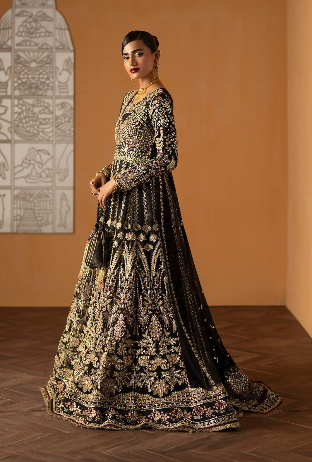 A woman models a long black Anarkali gown with intricate gold and beige embroidery, standing in a warm-toned room with a terracotta wall and wooden floor.