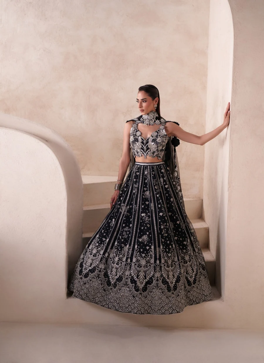 A woman models a black and silver heavily embroidered traditional Indian gown with a high-neck choker and flared skirt, posing on a beige stone staircase.