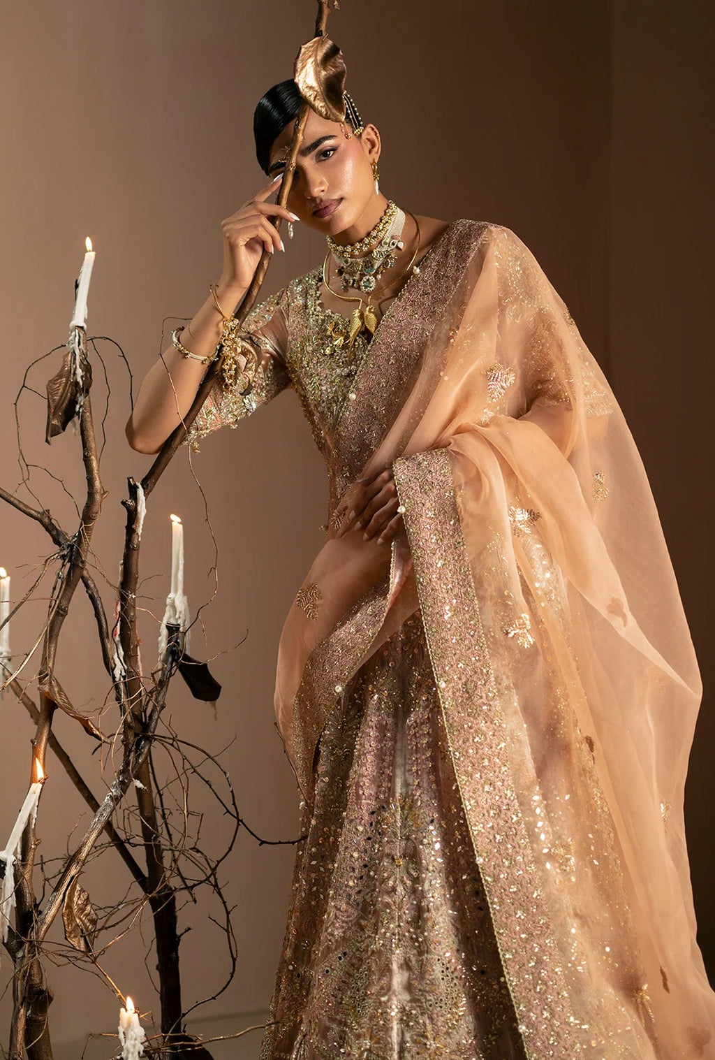 A woman models a heavily embellished peach-beige traditional outfit with a matching sheer dupatta, gold jewelry, and a decorative headpiece, posed beside a branch-like candle holder against a warm brown studio background.