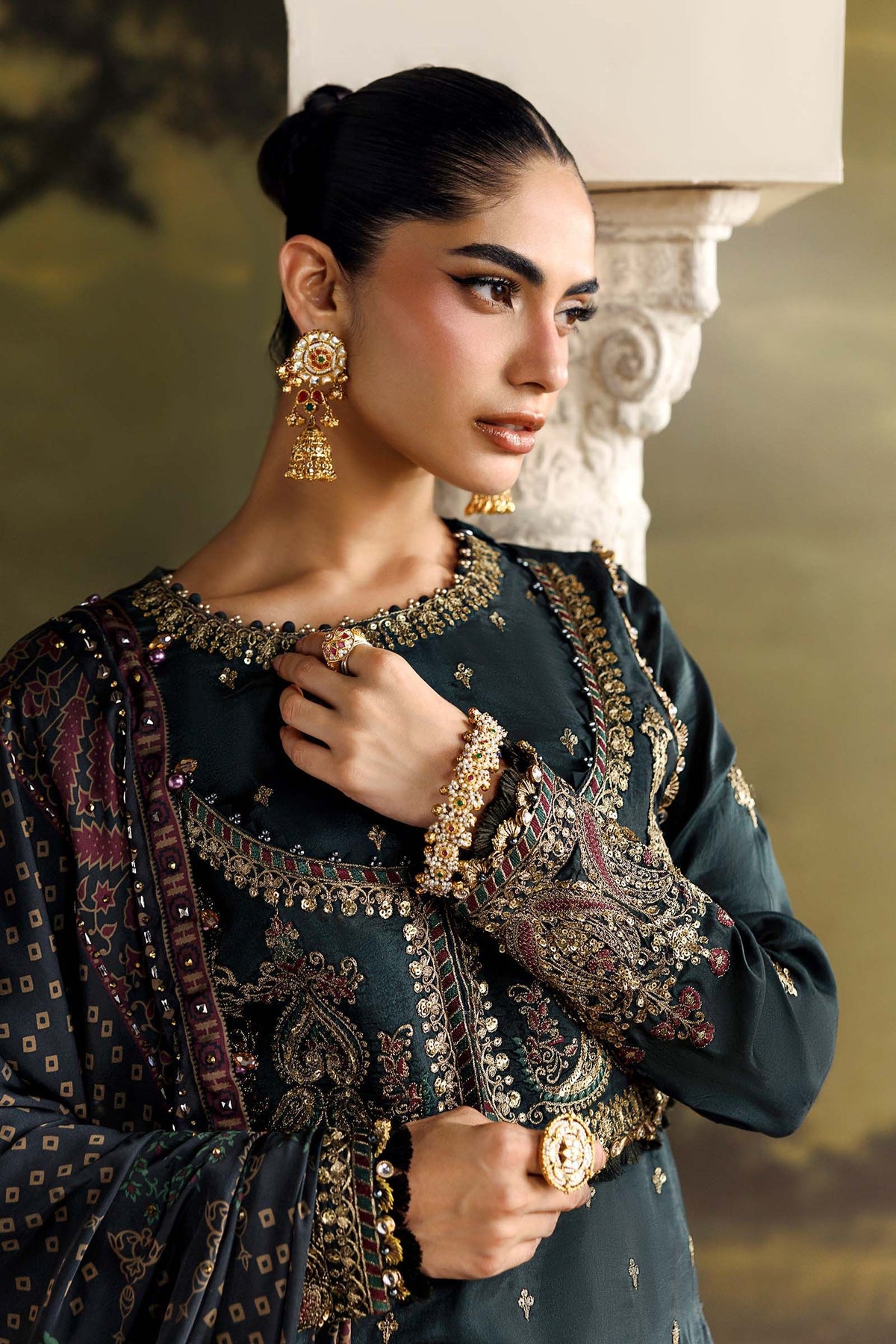 A woman models a dark teal green embroidered traditional outfit with intricate gold and multicolor detailing, styled with gold jewelry against a warm, classical background.