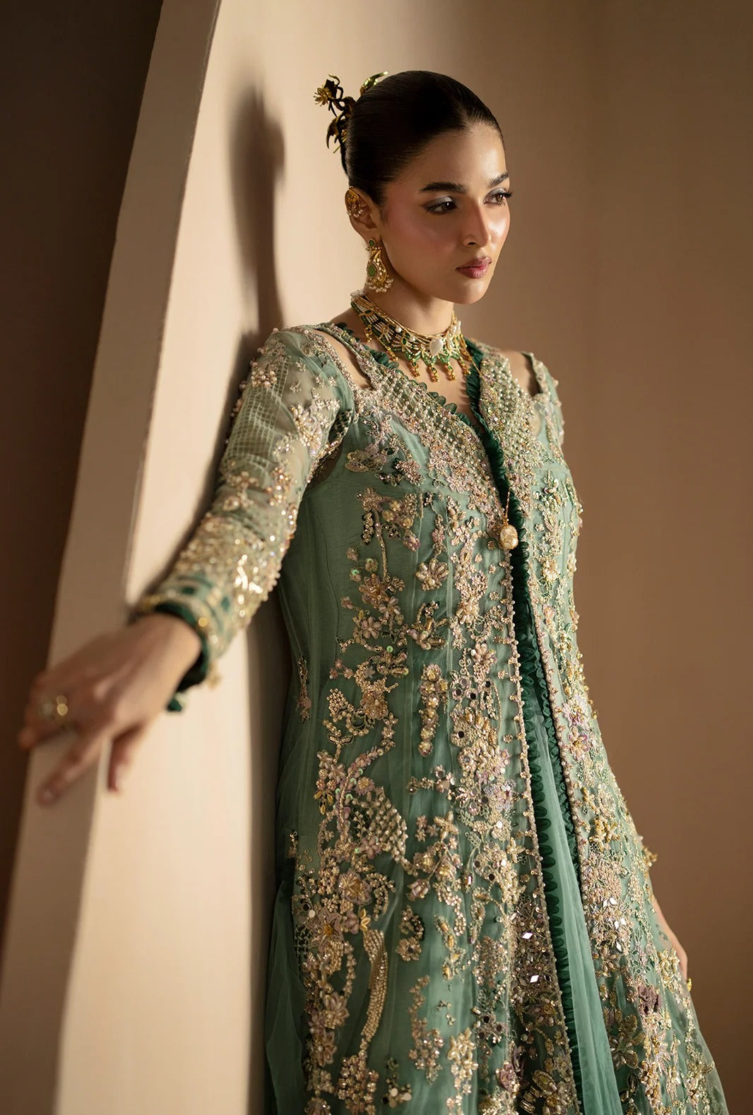 A woman models a long sage green anarkali-style dress with extensive gold and silver embroidery, standing against a warm beige wall.