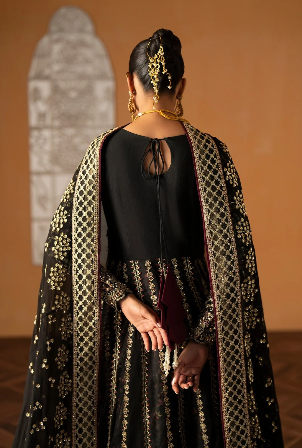 A woman shown from behind wearing a black traditional outfit with an ornate gold embroidered outer layer and matching jewelry, standing against a warm brown wall.