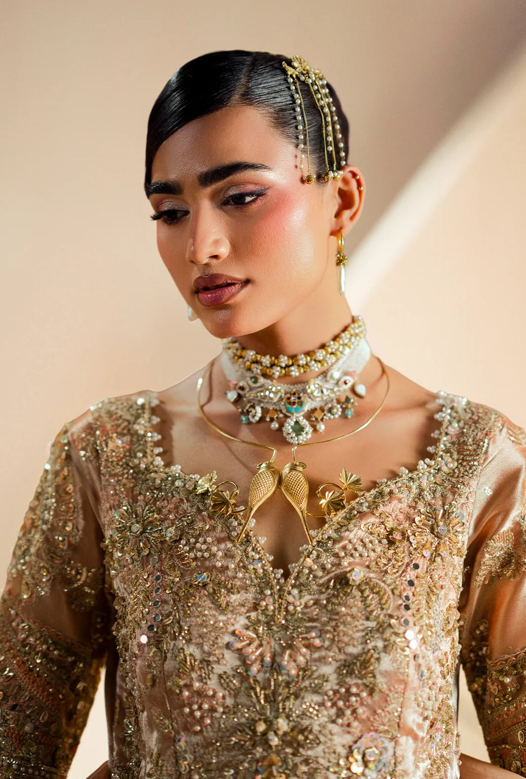 A close-up of a model wearing an ornate gold and beige embellished traditional outfit with layered gold necklaces and a matching headpiece against a soft beige background.