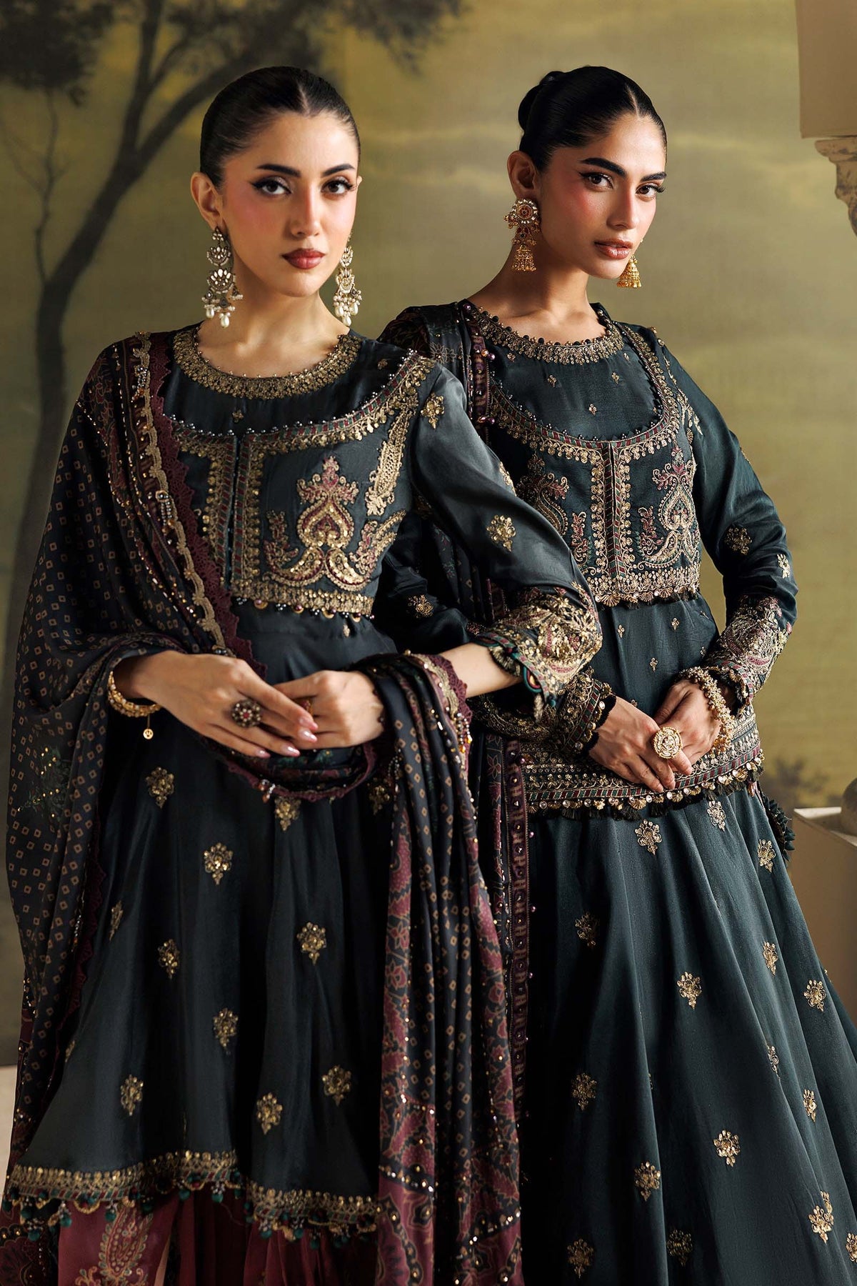 Two women modeling matching dark teal-green traditional South Asian outfits with heavy gold embroidery, patterned dupattas, and ornate jewelry against a warm beige background.