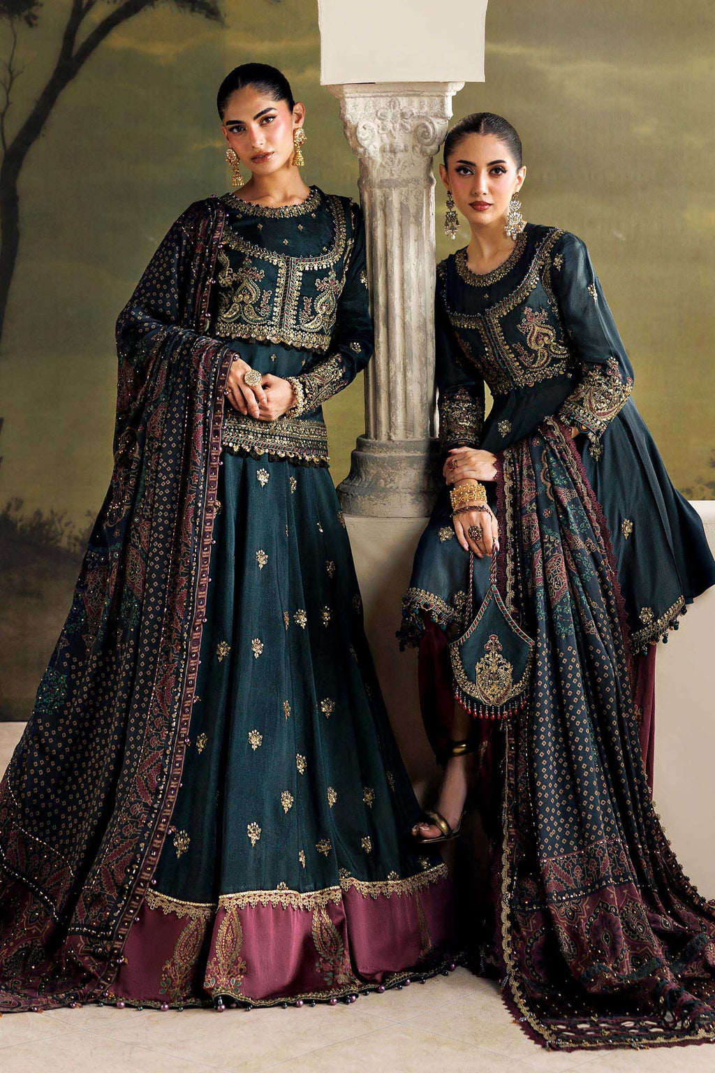 Two women modeling traditional dark teal Anarkali suits with gold embroidery and deep maroon borders, posed in front of a classical stone column and painted backdrop.
