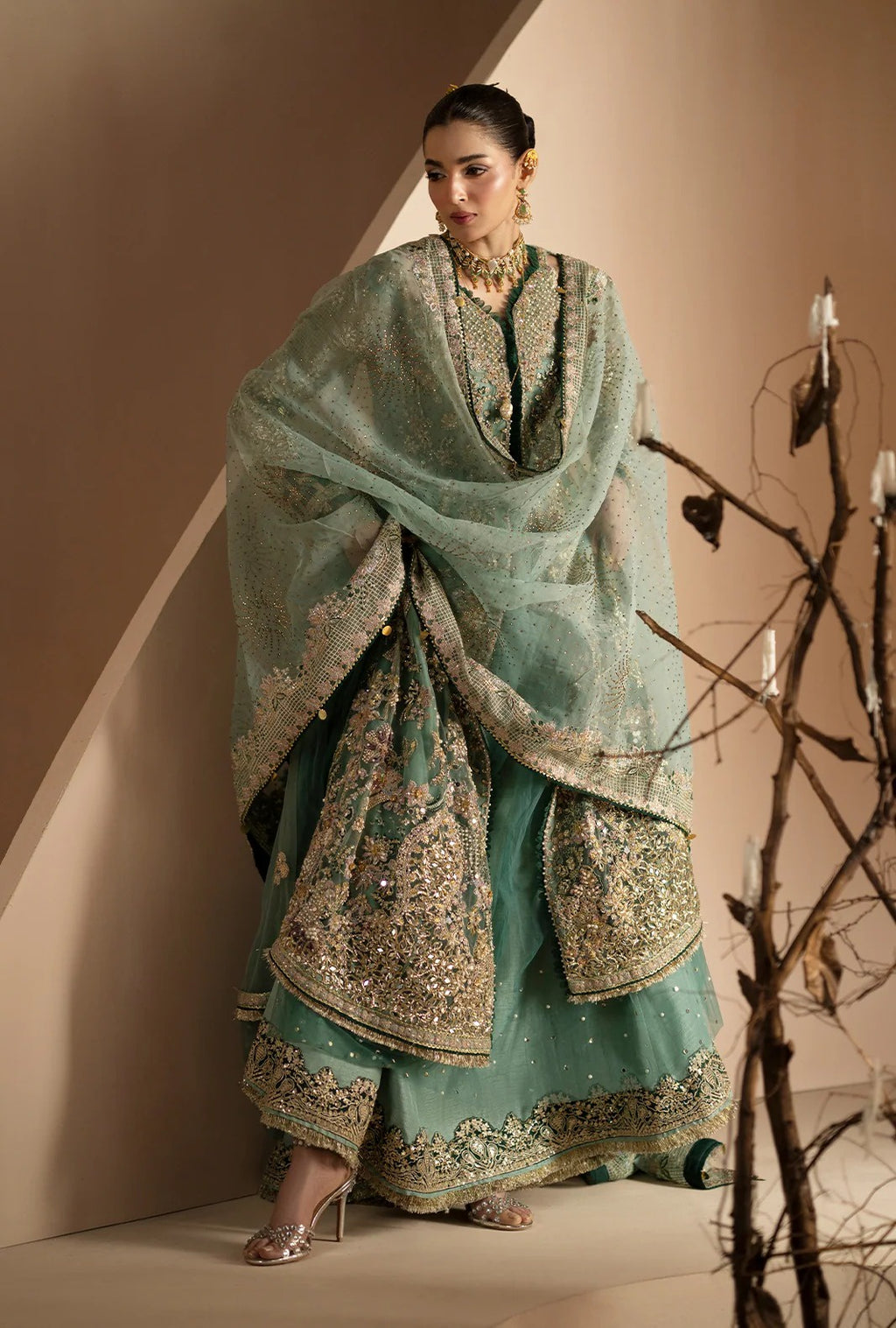 A woman models a traditional South Asian outfit consisting of a mint green embroidered kurta, matching trousers, and a sheer dupatta with gold embroidery, set against a minimalist beige background with decorative branches.