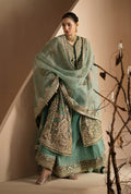 A woman models a traditional South Asian outfit consisting of a mint green embroidered kurta, matching trousers, and a sheer dupatta with gold embroidery, set against a minimalist beige background with decorative branches.
