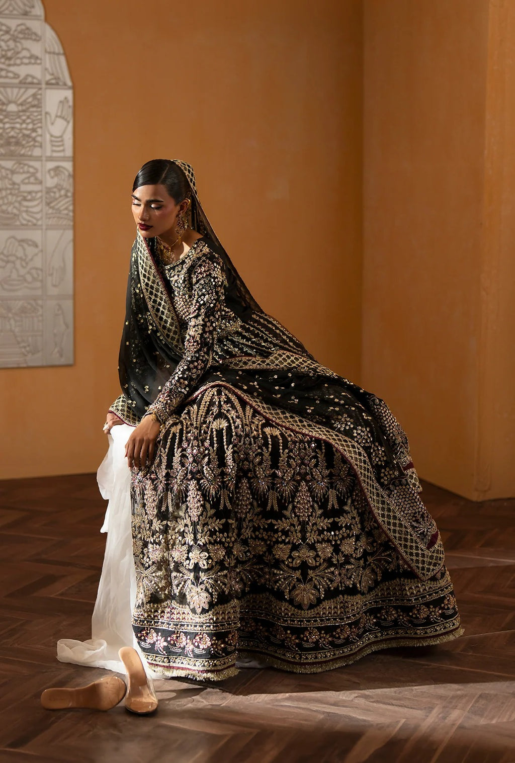 A woman wearing an ornate black and gold embroidered traditional outfit with a long veil, seated on a wooden floor against a warm tan wall. A white dupatta is draped beside her, and beige shoes are placed on the floor.