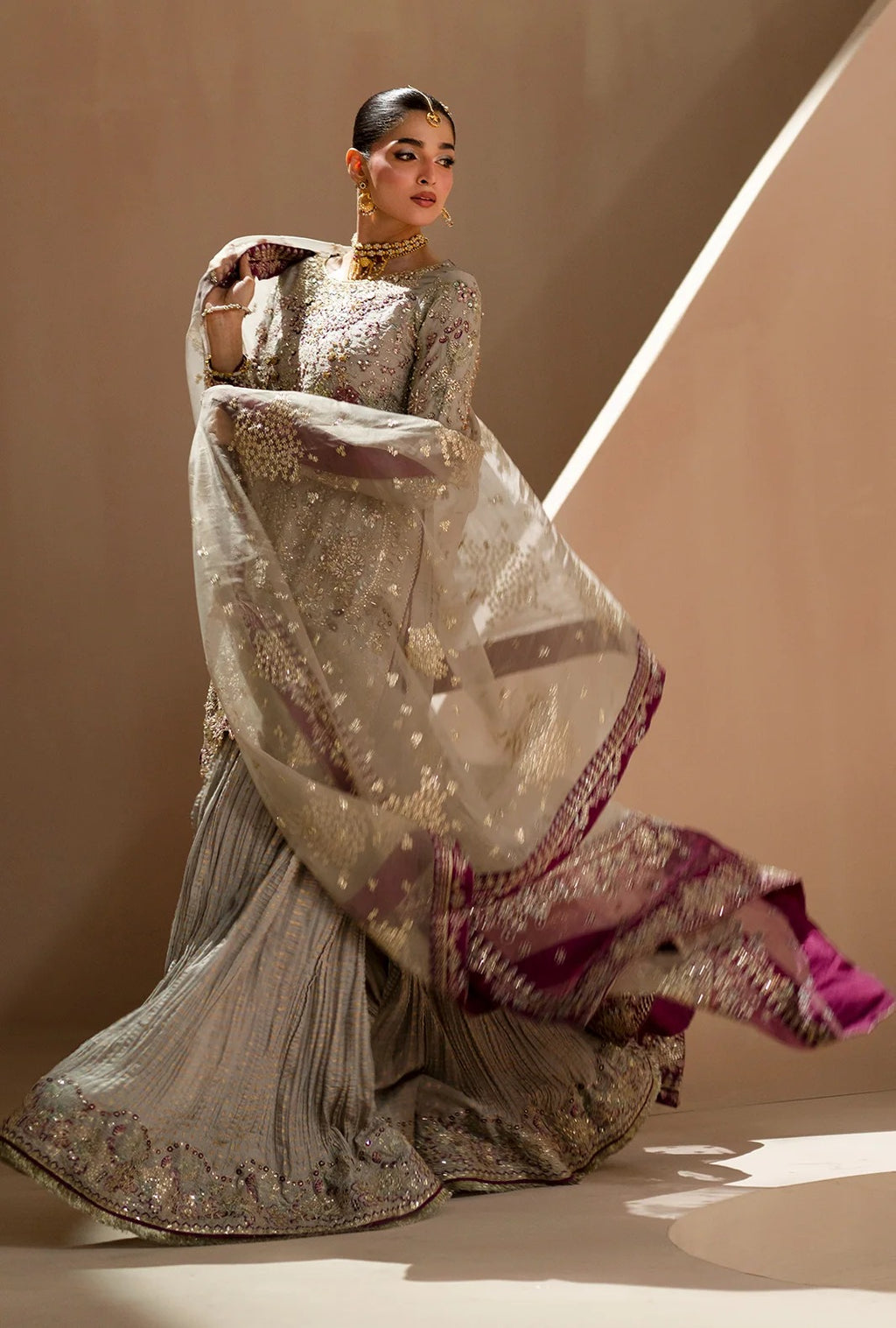 A model wearing a heavily embroidered traditional South Asian outfit, featuring a silver-gray pleated skirt, a matching long-sleeve embroidered blouse, and a sheer dupatta with gold motifs and a deep magenta border, posed against a warm beige studio backdrop.