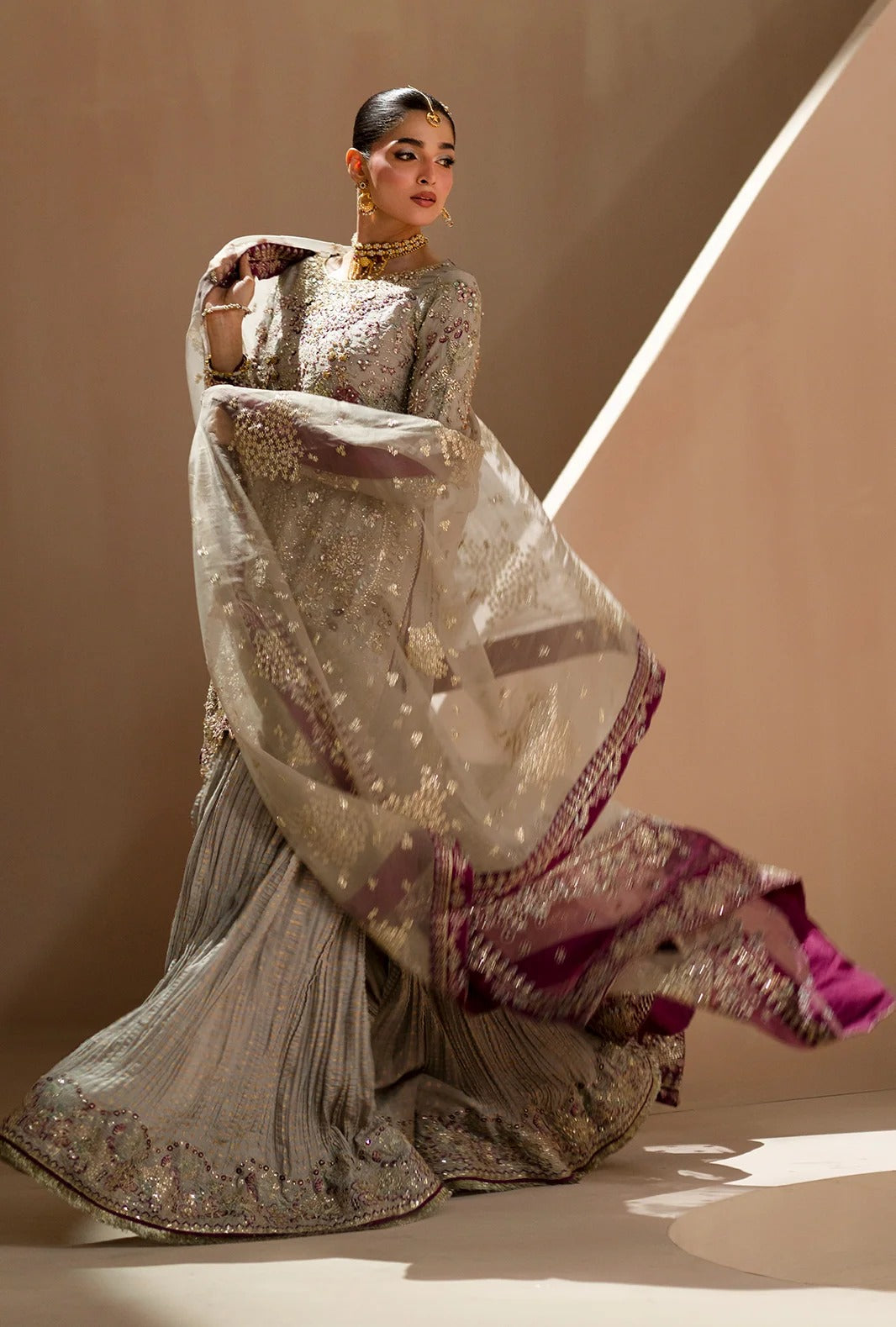 A model wearing a heavily embroidered traditional South Asian outfit, featuring a silver-gray pleated skirt, a matching long-sleeve embroidered blouse, and a sheer dupatta with gold motifs and a deep magenta border, posed against a warm beige studio backdrop.