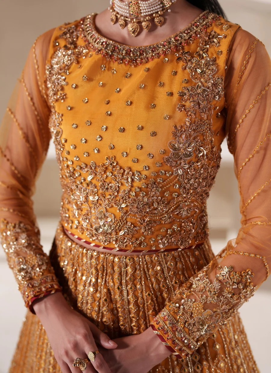 A close-up of a model wearing a traditional Indian outfit featuring a mustard yellow embroidered blouse with intricate gold metallic embroidery, sequins, and mirror work, paired with a matching pleated skirt.