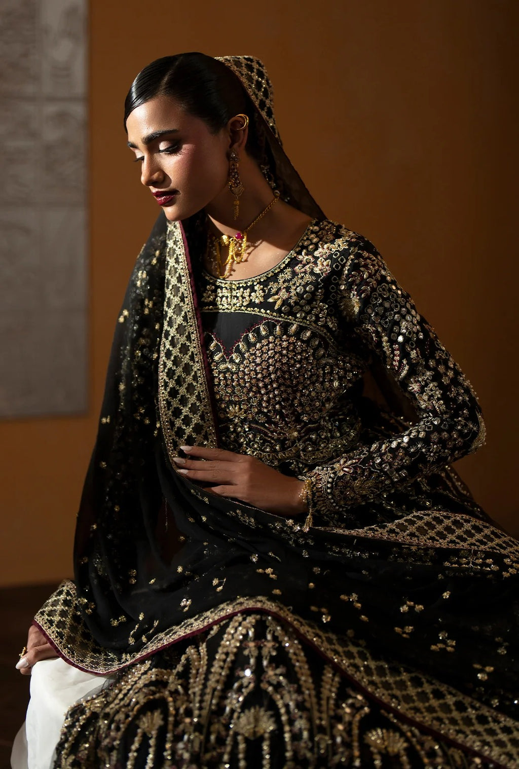 A woman wearing an ornate black traditional outfit with extensive gold embroidery and beadwork, seated against a warm brown background.