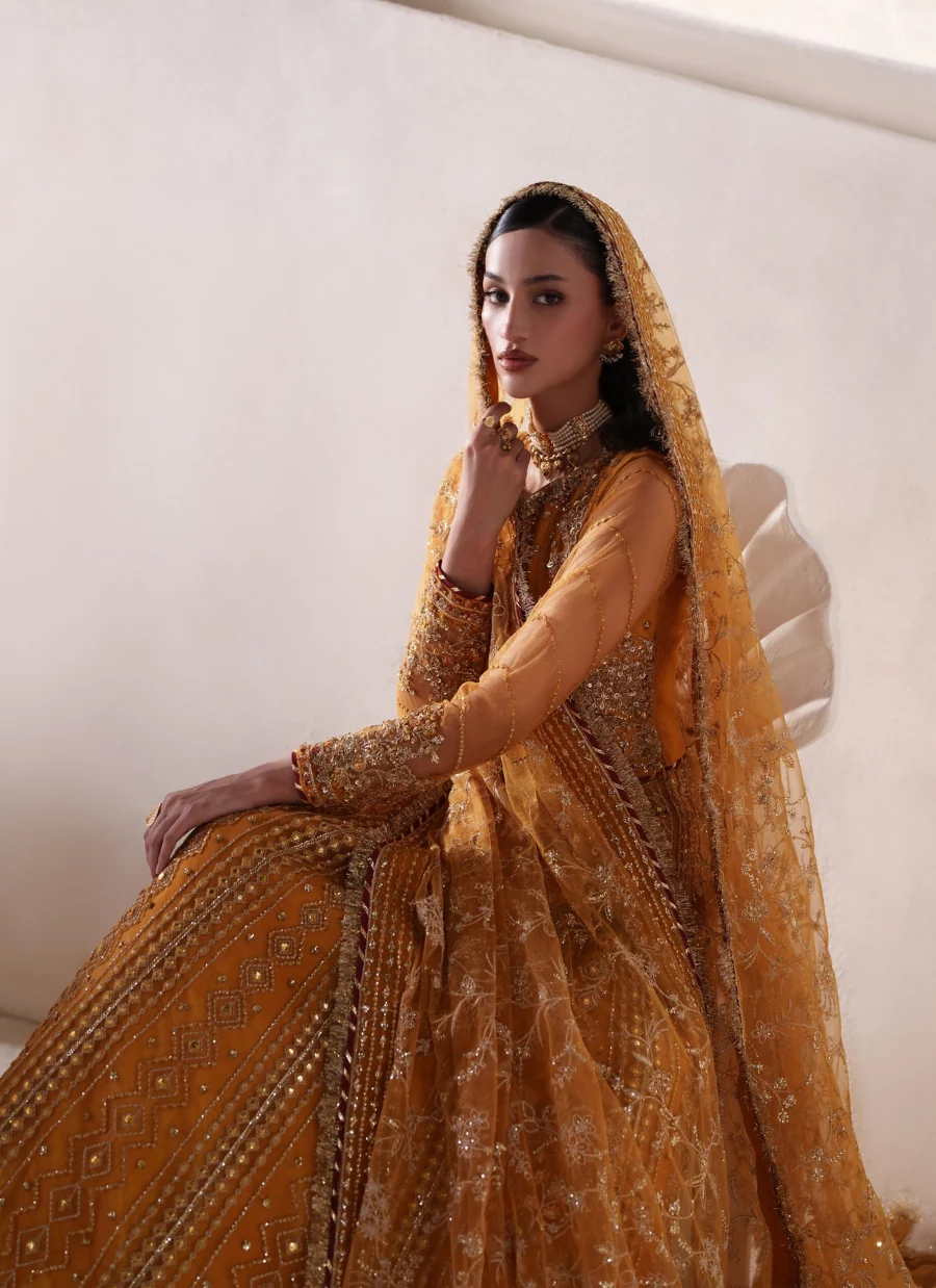 A woman wearing an ornate mustard yellow and gold embroidered traditional outfit with a matching sheer veil, posed against a light beige wall.