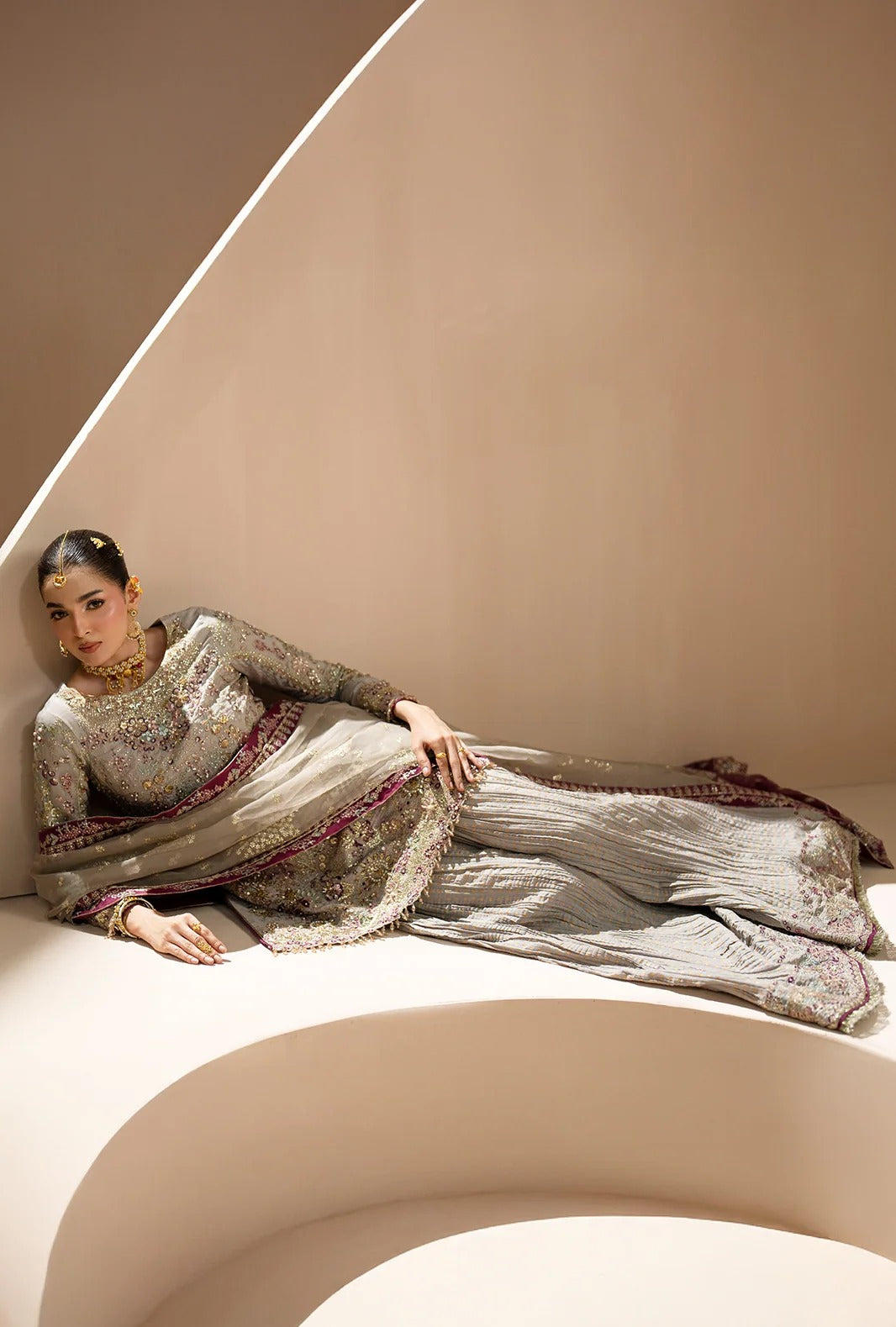 A model reclines on a curved beige platform wearing a heavily embroidered light gray and silver traditional outfit with a matching dupatta and gold jewelry.