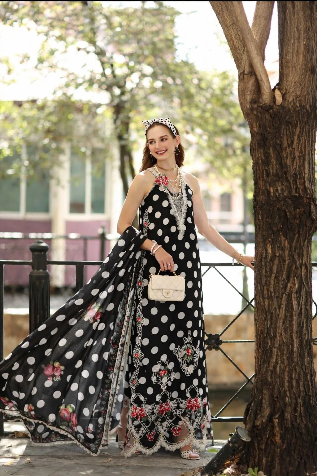 Woman in a black and white polka dot dress with floral details standing next to a tree.