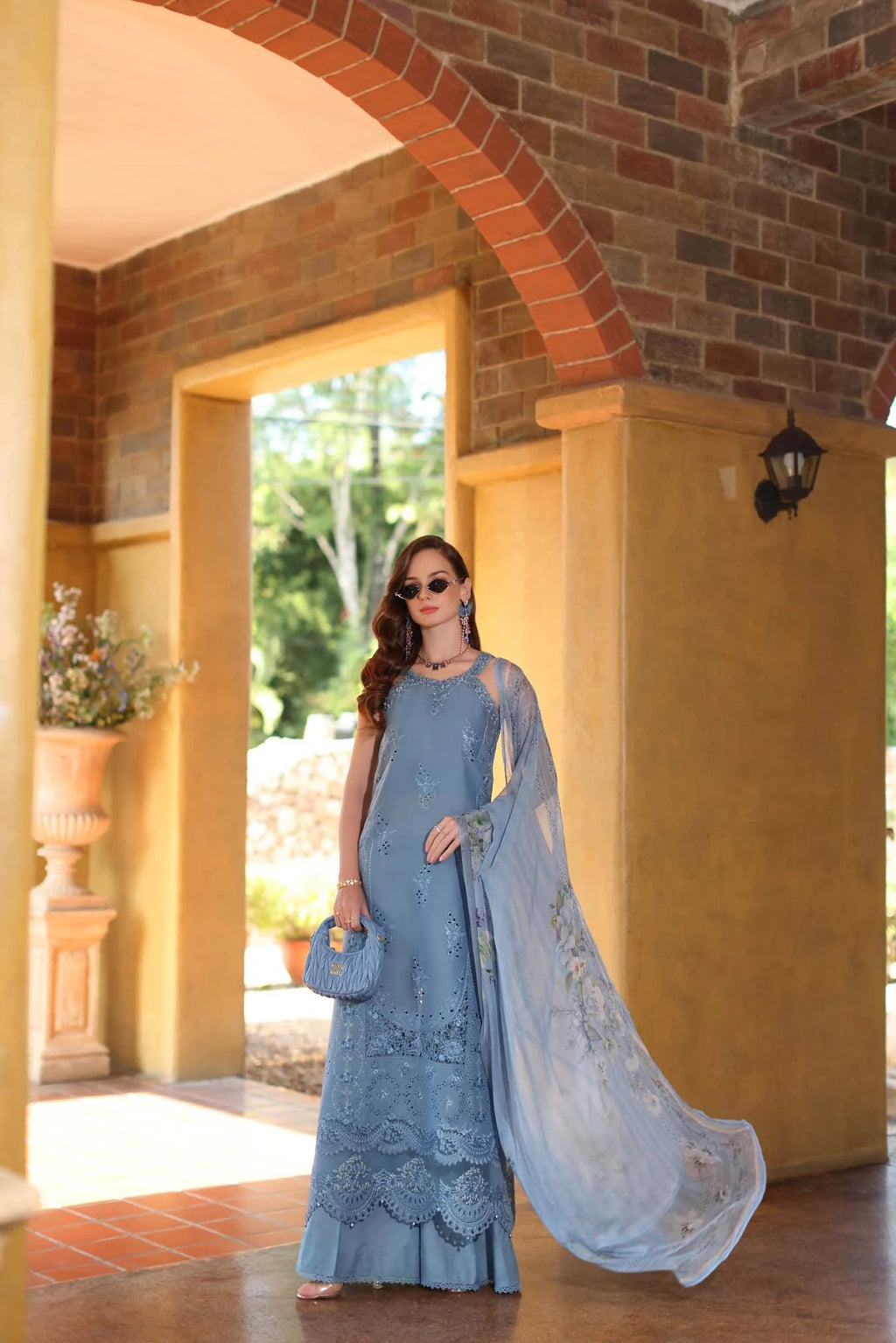 Woman in a blue lace dress standing in a sunlit outdoor setting with arches and plants.
