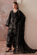Woman wearing a black embroidered outfit with a long scarf against a light wall.
