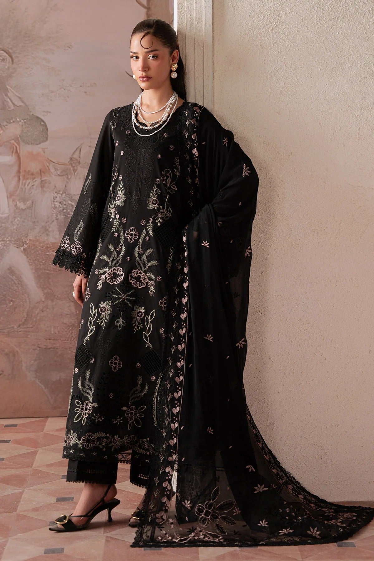 Woman wearing a black embroidered outfit with a long scarf against a light wall.