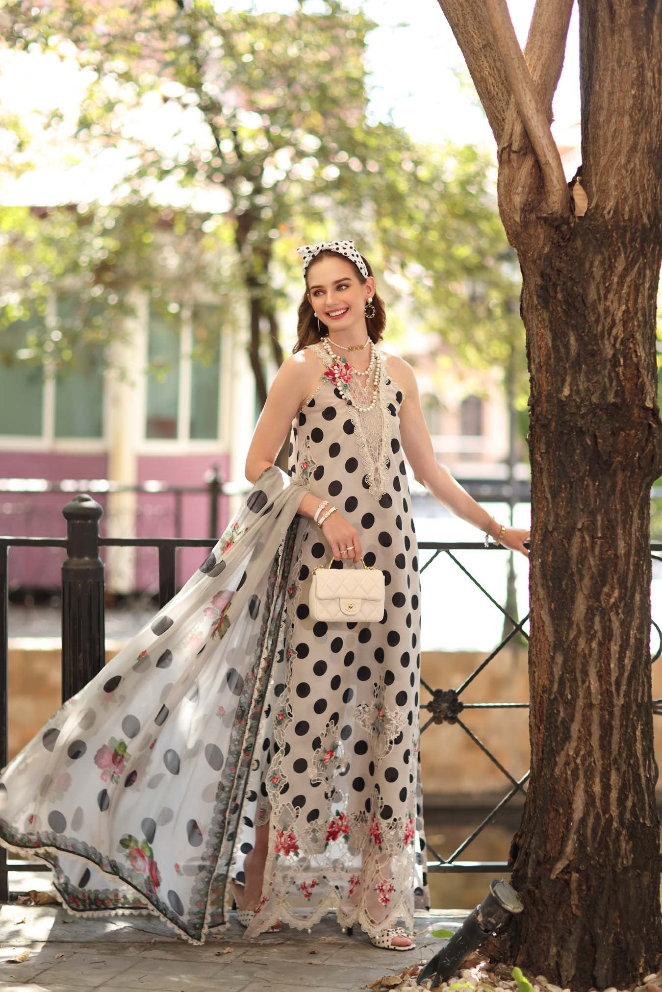 Woman in a polka dot dress with a floral headband and handbag standing next to a tree.