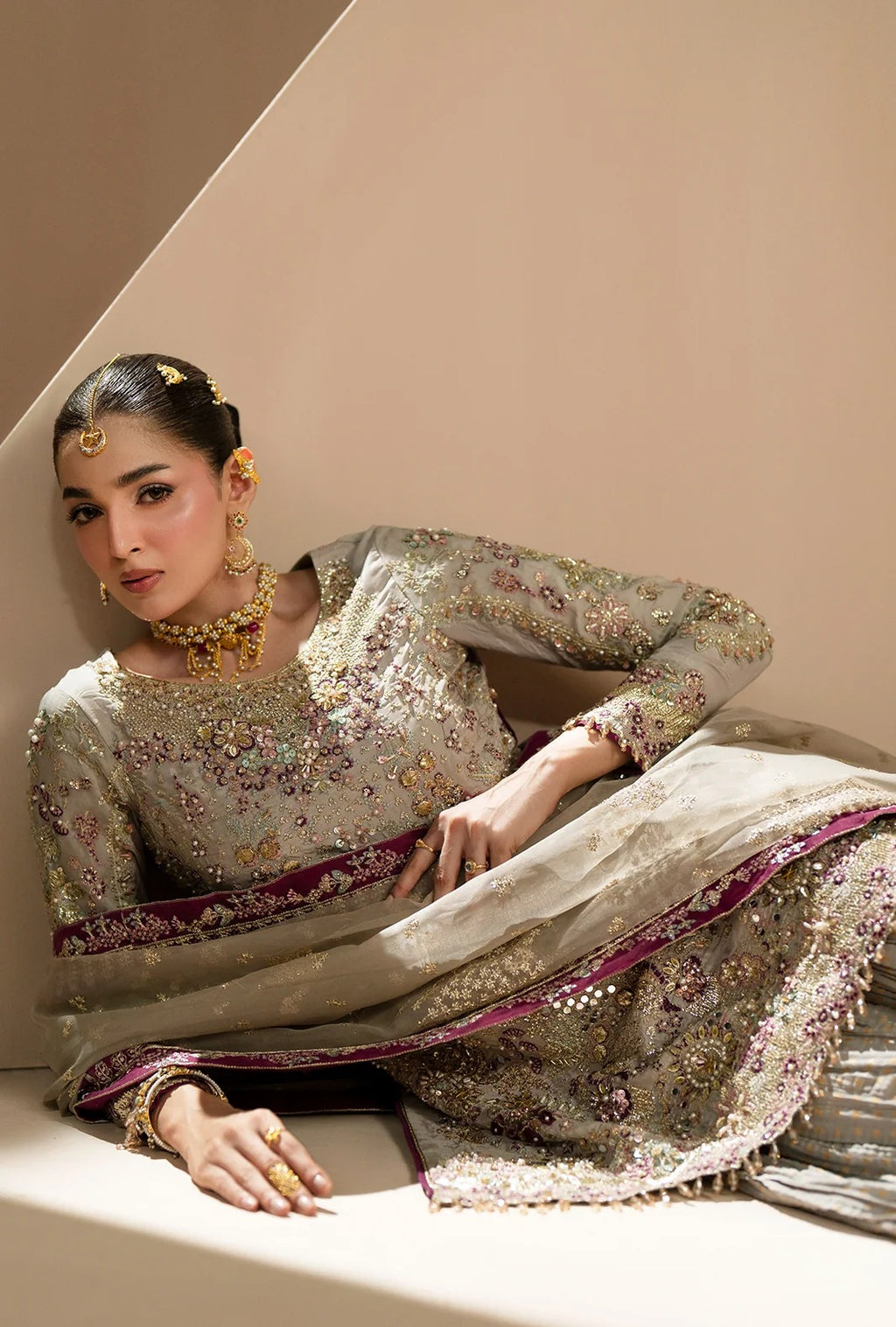 A woman models a traditional Indian outfit consisting of a heavily embroidered beige and silver kurta with a matching dupatta draped over her lap, set against a minimalist beige background.
