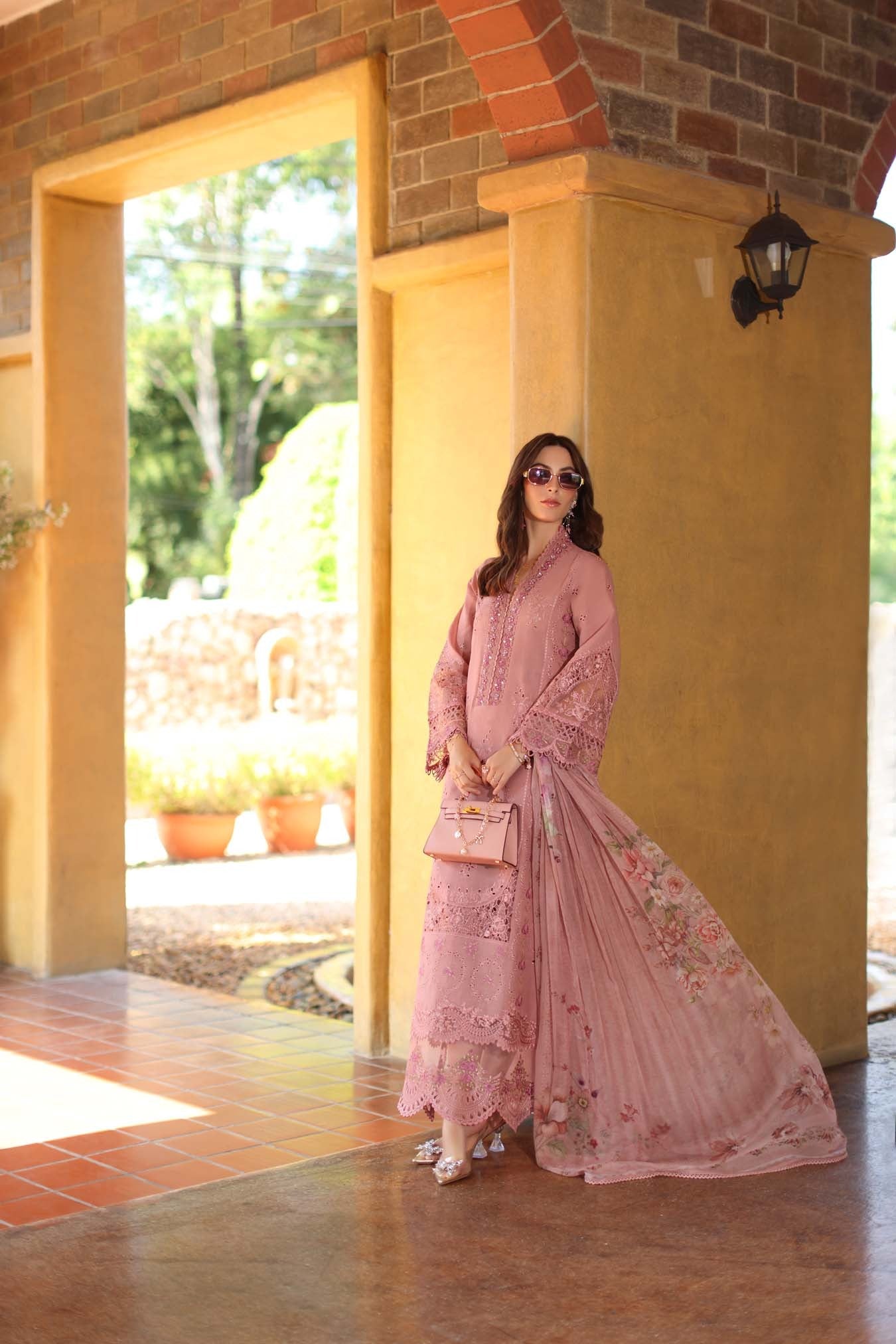 Woman in a pink traditional outfit standing in a sunlit outdoor setting with a yellow wall.