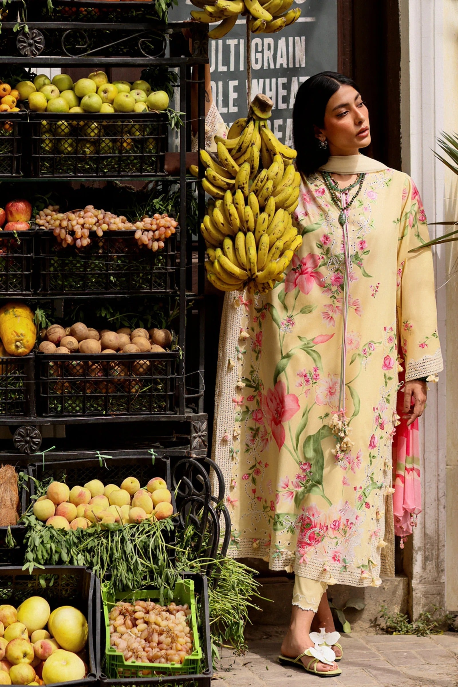 Woman in a floral coat with bananas over her shoulder in front of a fruit stand.