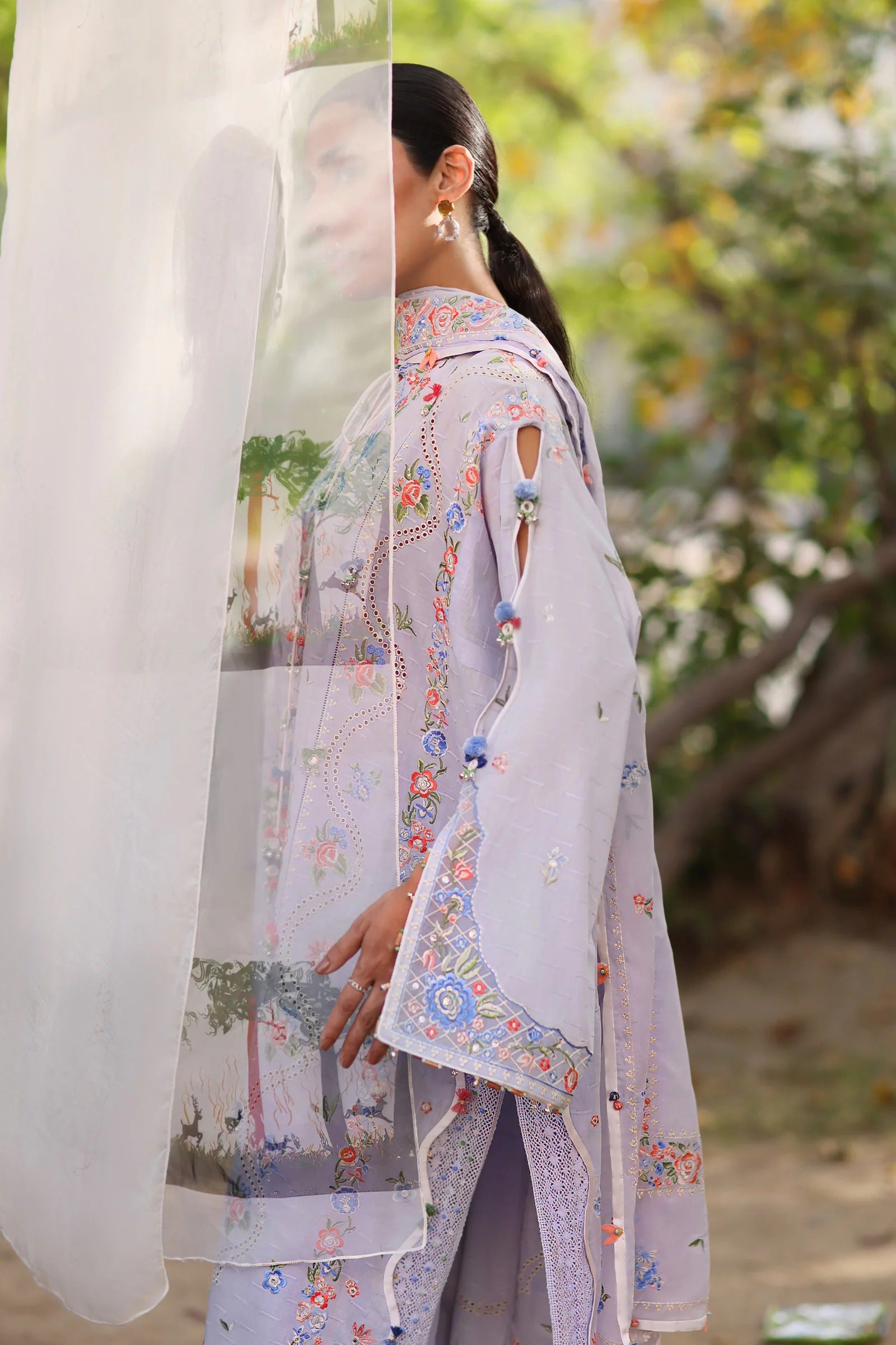 Woman wearing a light-colored traditional outfit with floral patterns, standing outdoors with greenery in the background.