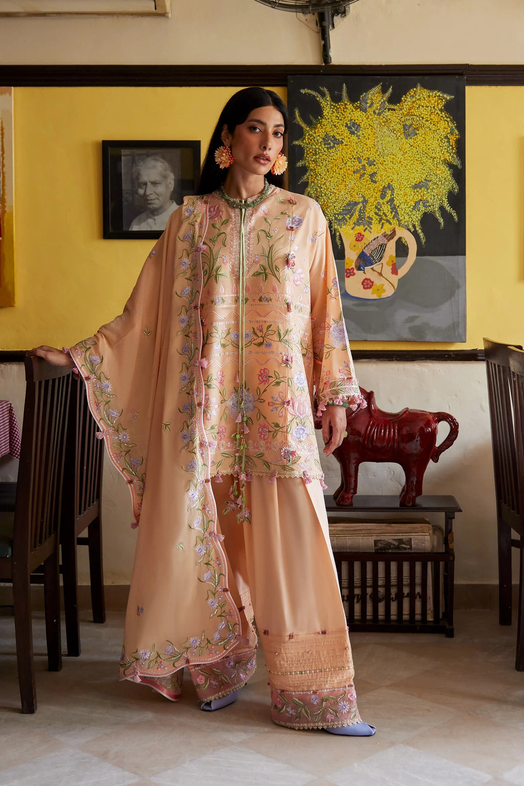 Woman in a peach floral traditional outfit standing in a room with yellow walls and decorative items.