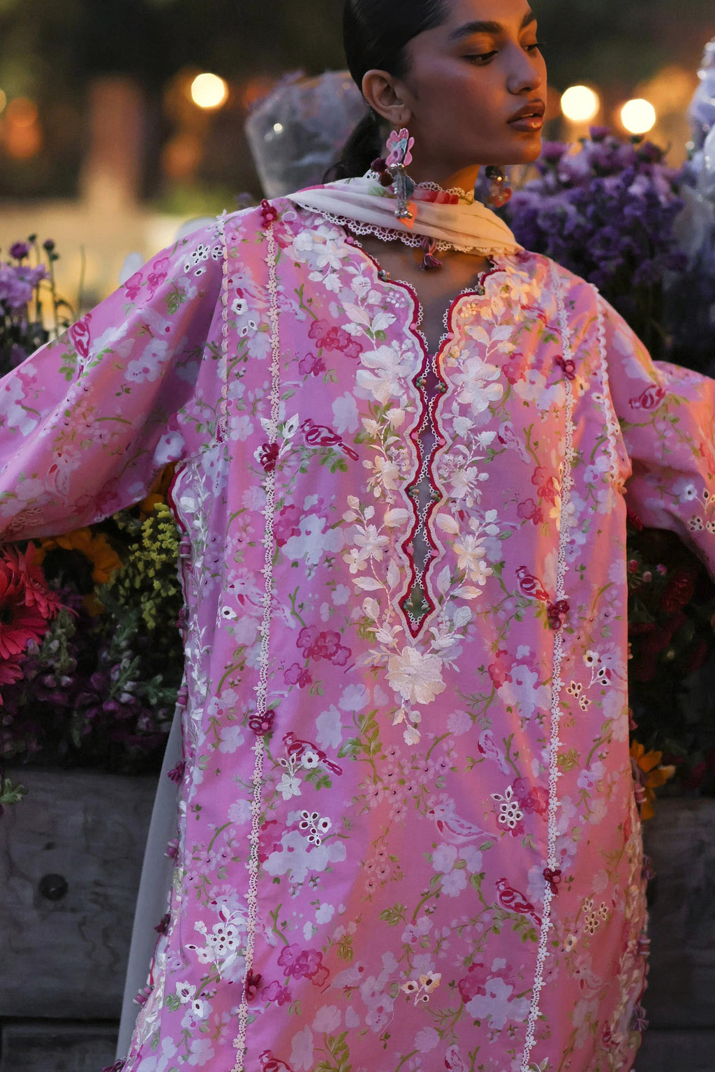 Woman wearing a pink floral embroidered traditional outfit with a blurred background of flowers and lights.
