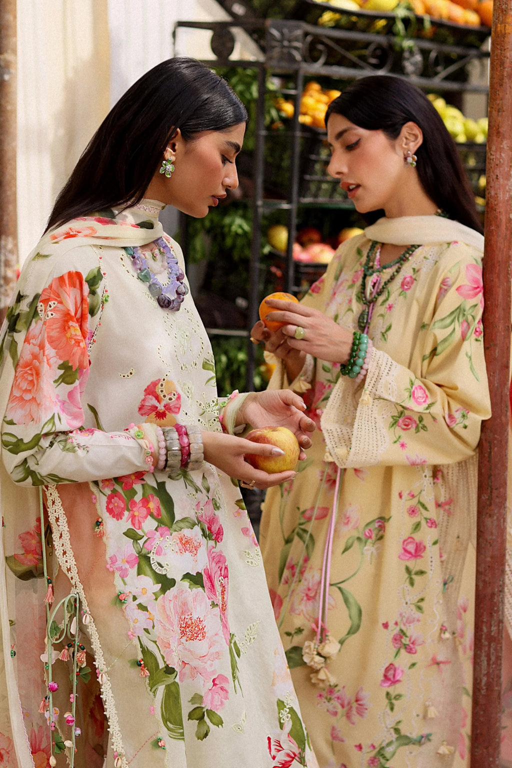 Two women in floral dresses standing next to each other, holding fruits.
