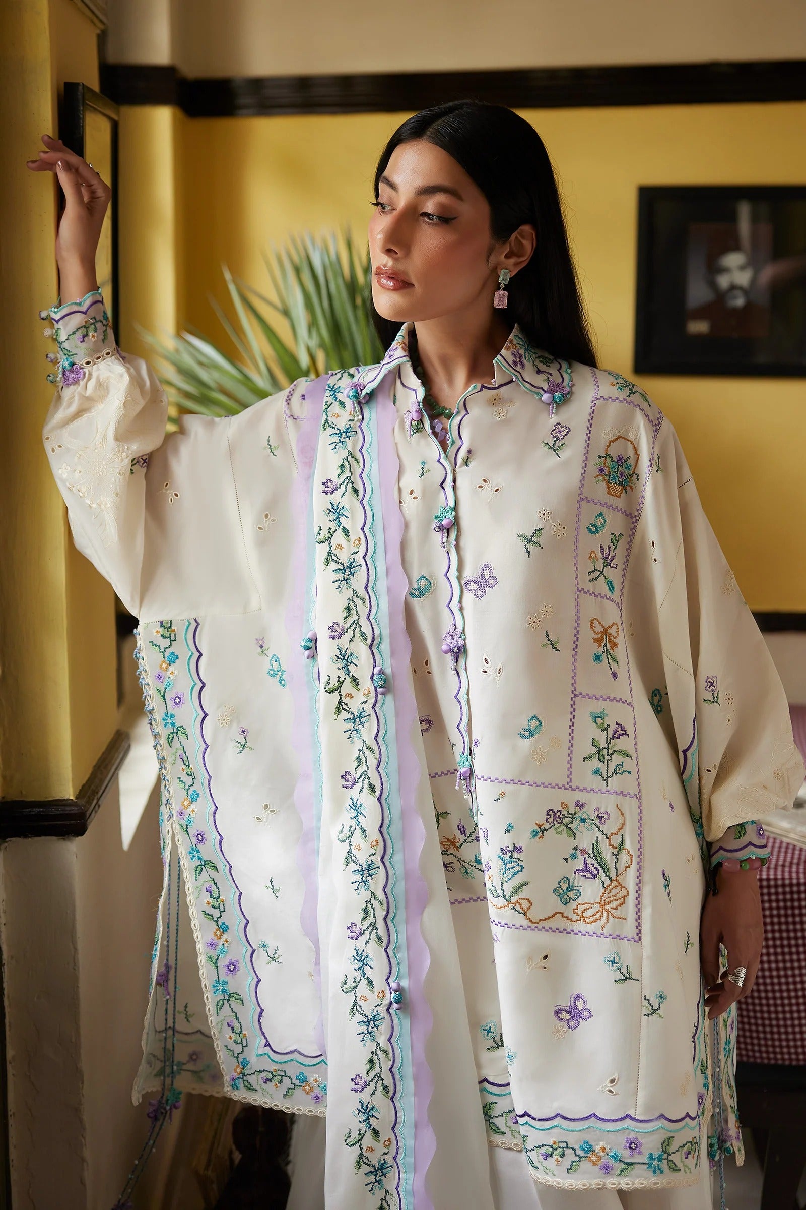 Woman wearing a traditional embroidered outfit in a room with yellow walls and a plant.