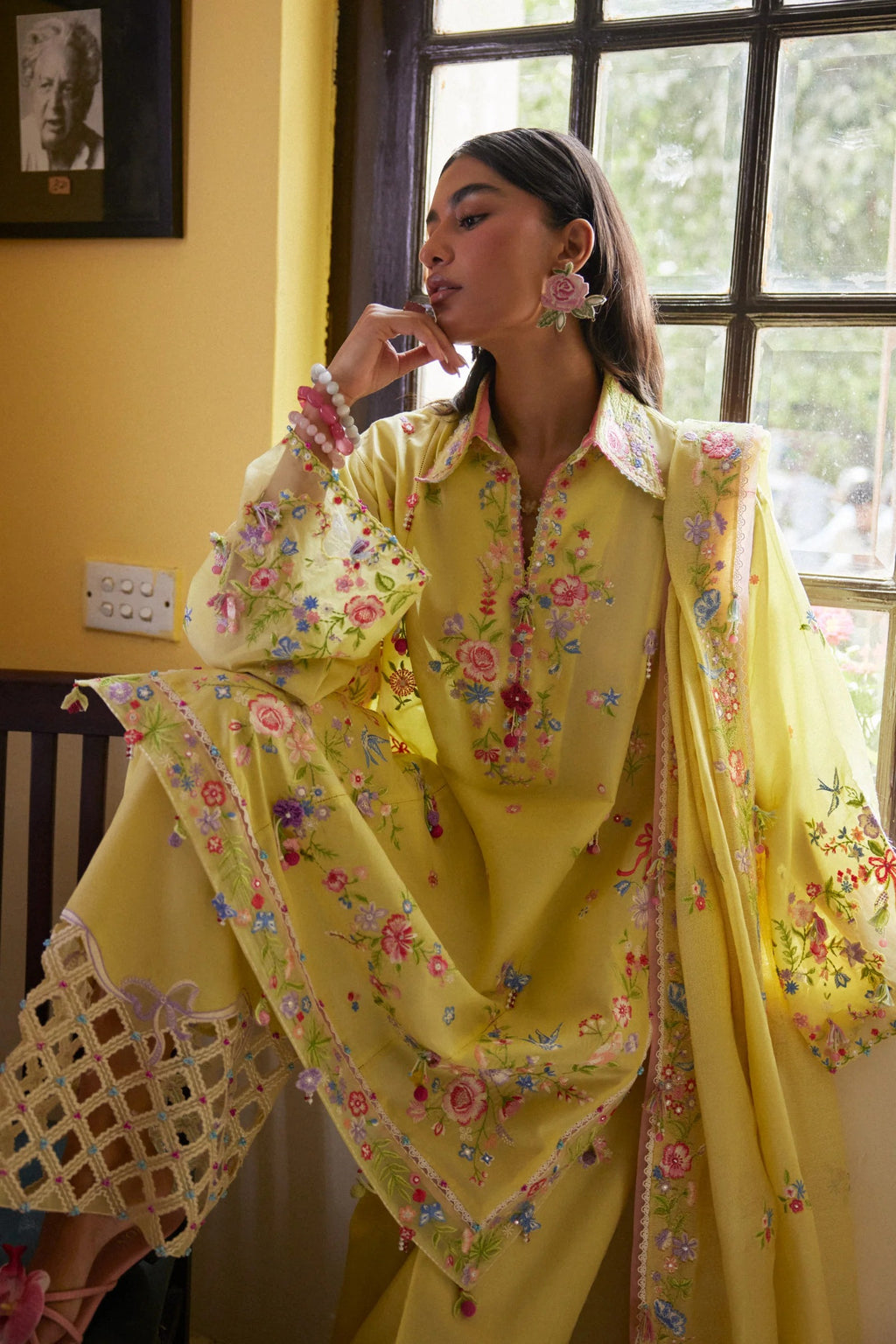 Woman in a yellow floral outfit standing indoors with a window and wall decor in the background.