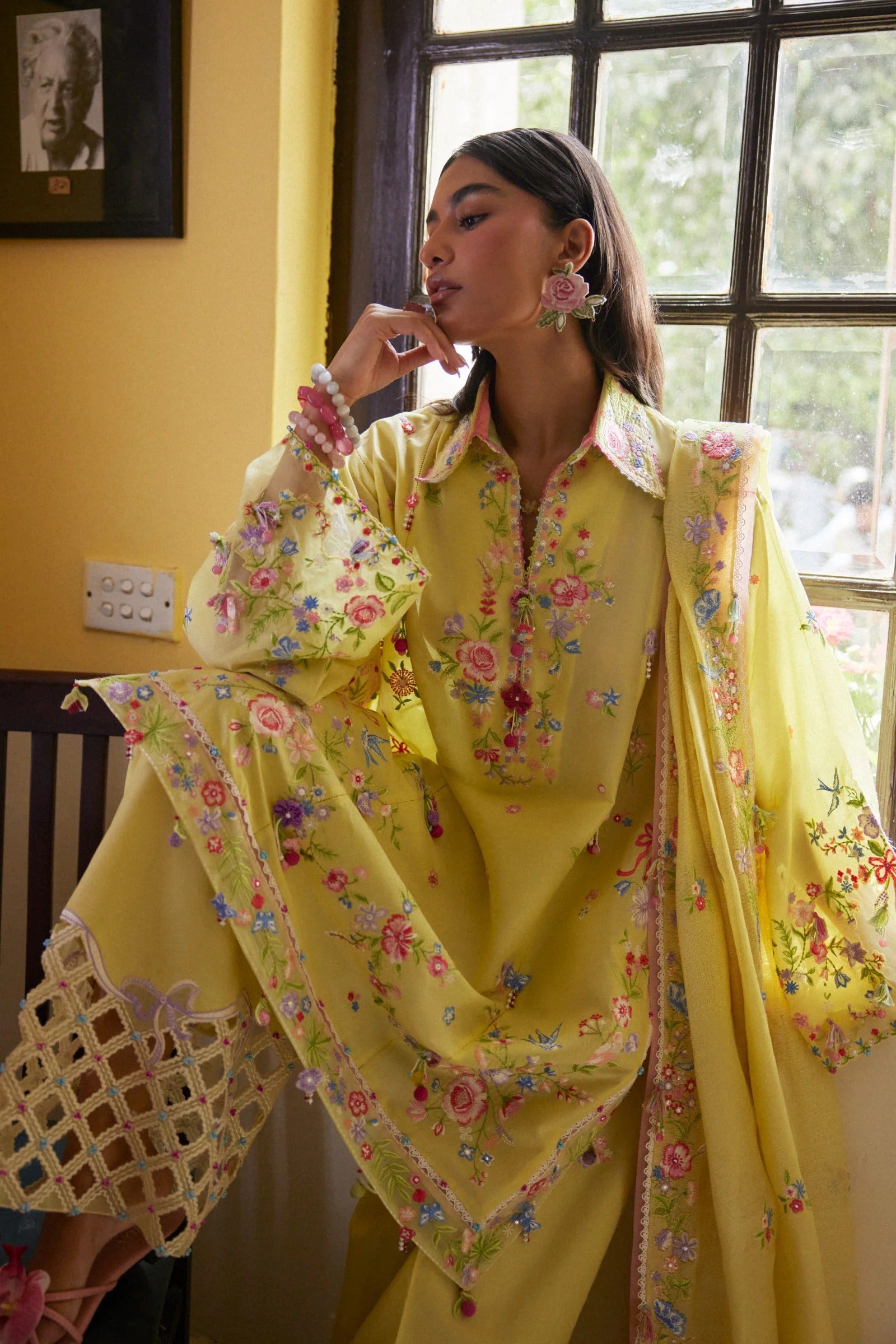 Woman in a yellow floral outfit standing indoors with a window and wall decor in the background.