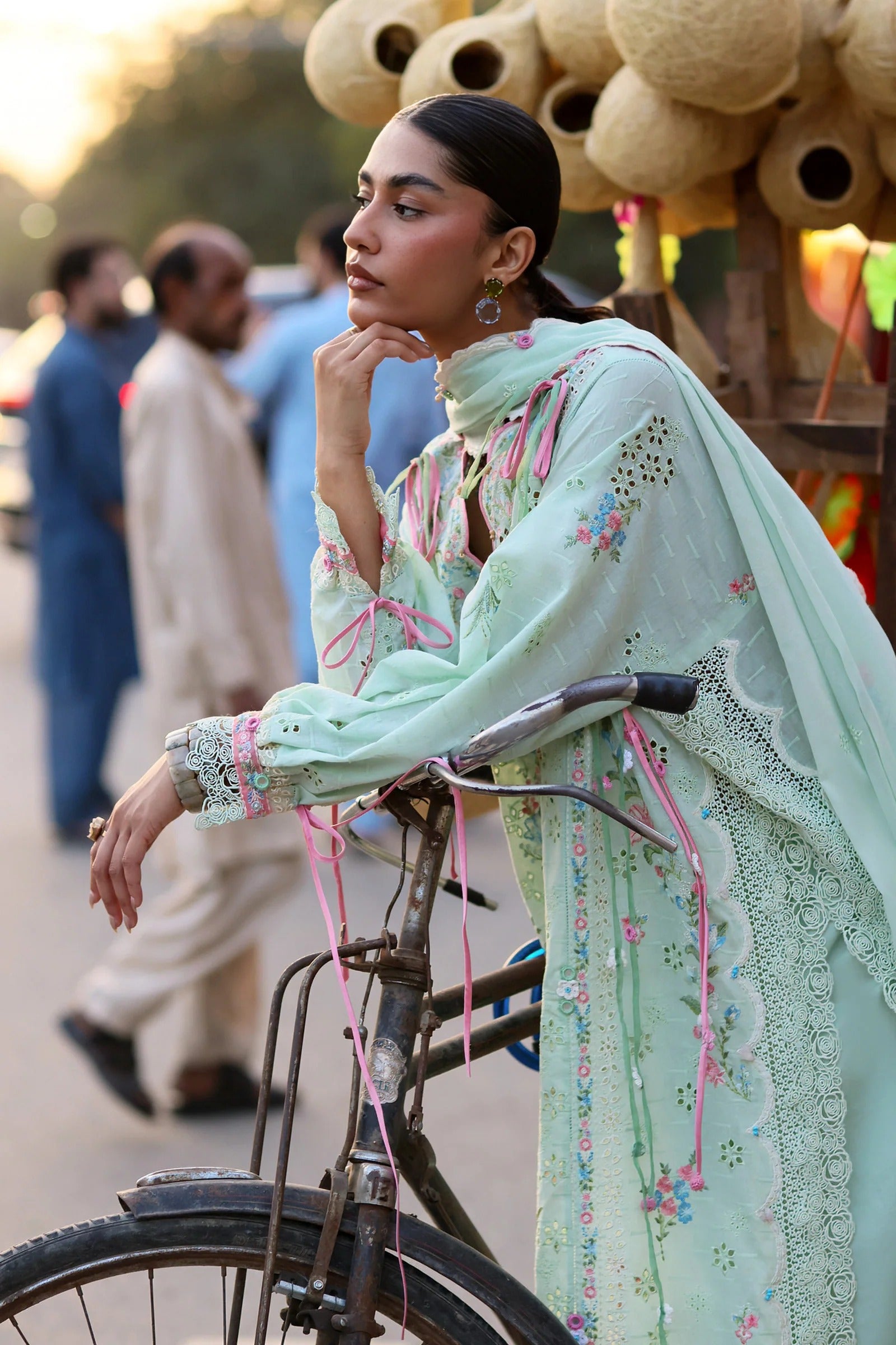Woman in a light green traditional outfit sitting on a bicycle with people and decorative elements in the background.