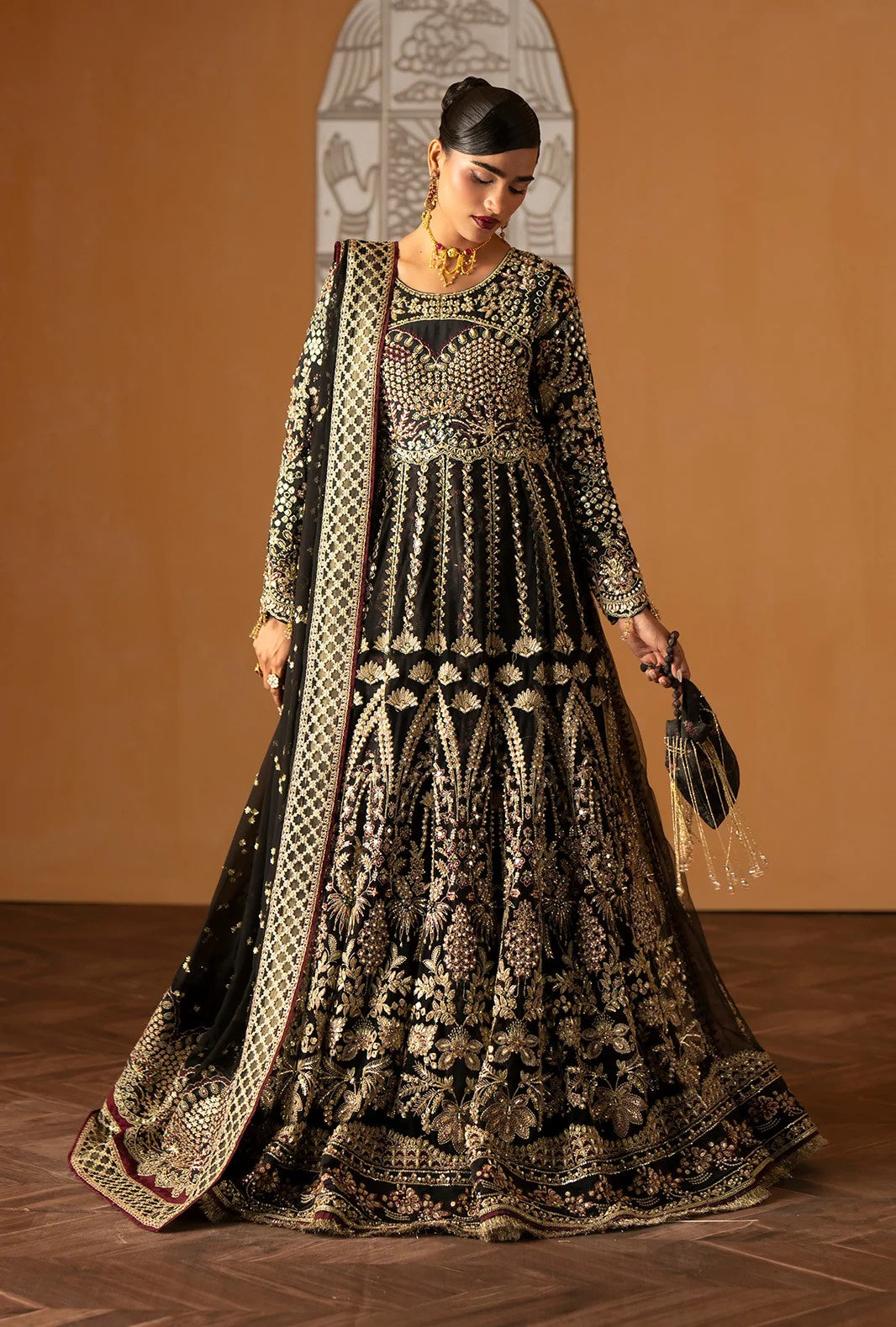 A woman models an ornate black and gold embroidered traditional gown with a matching dupatta in a warm indoor setting with a terracotta-colored wall.