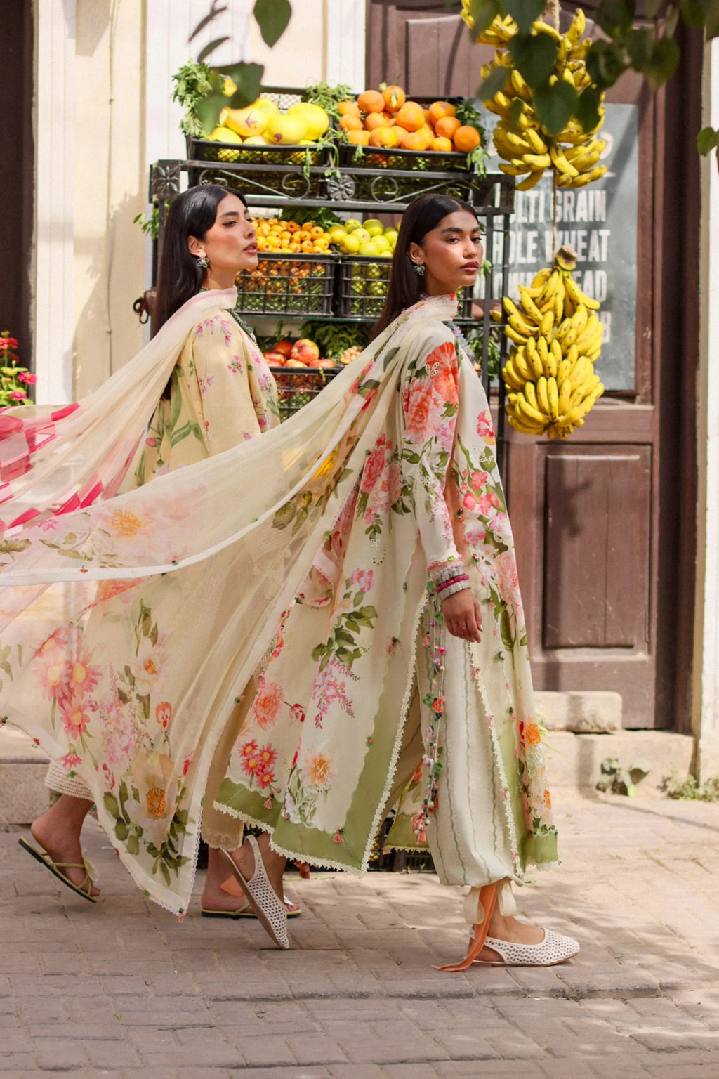 Two women in floral dresses walking past a fruit stand.