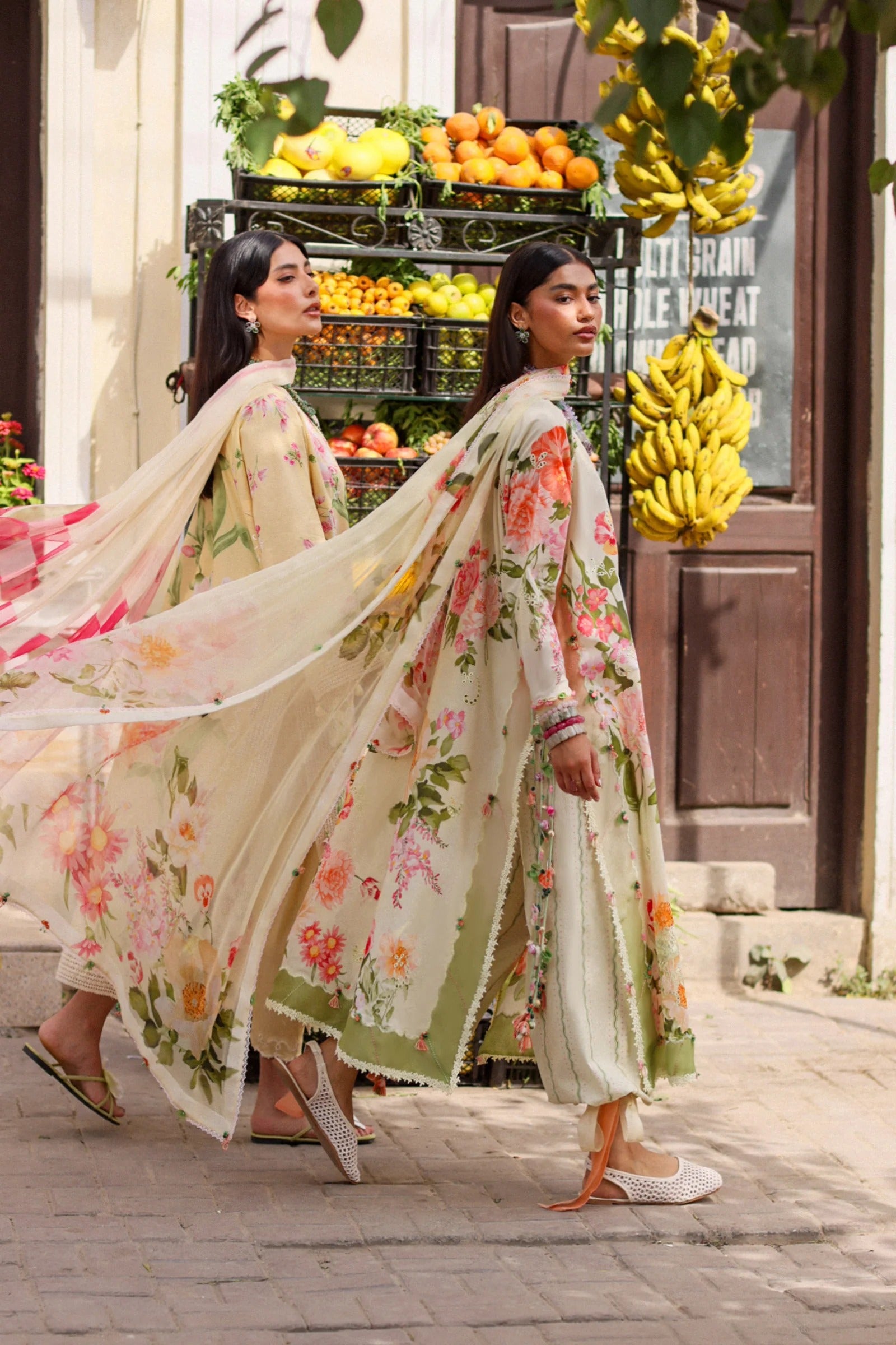 Two women in floral dresses walking past a fruit stand.
