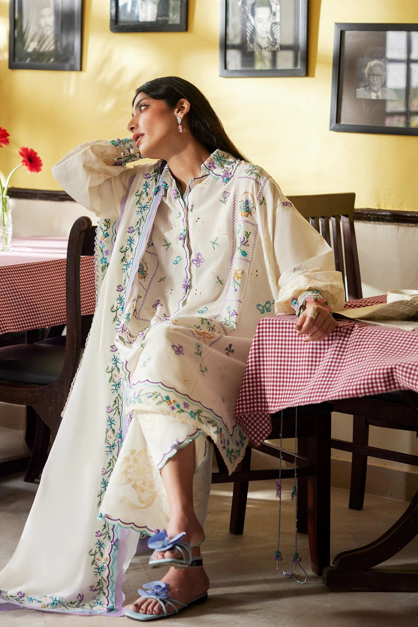 Woman in a white embroidered outfit sitting at a table with a yellow wall and framed pictures in the background.