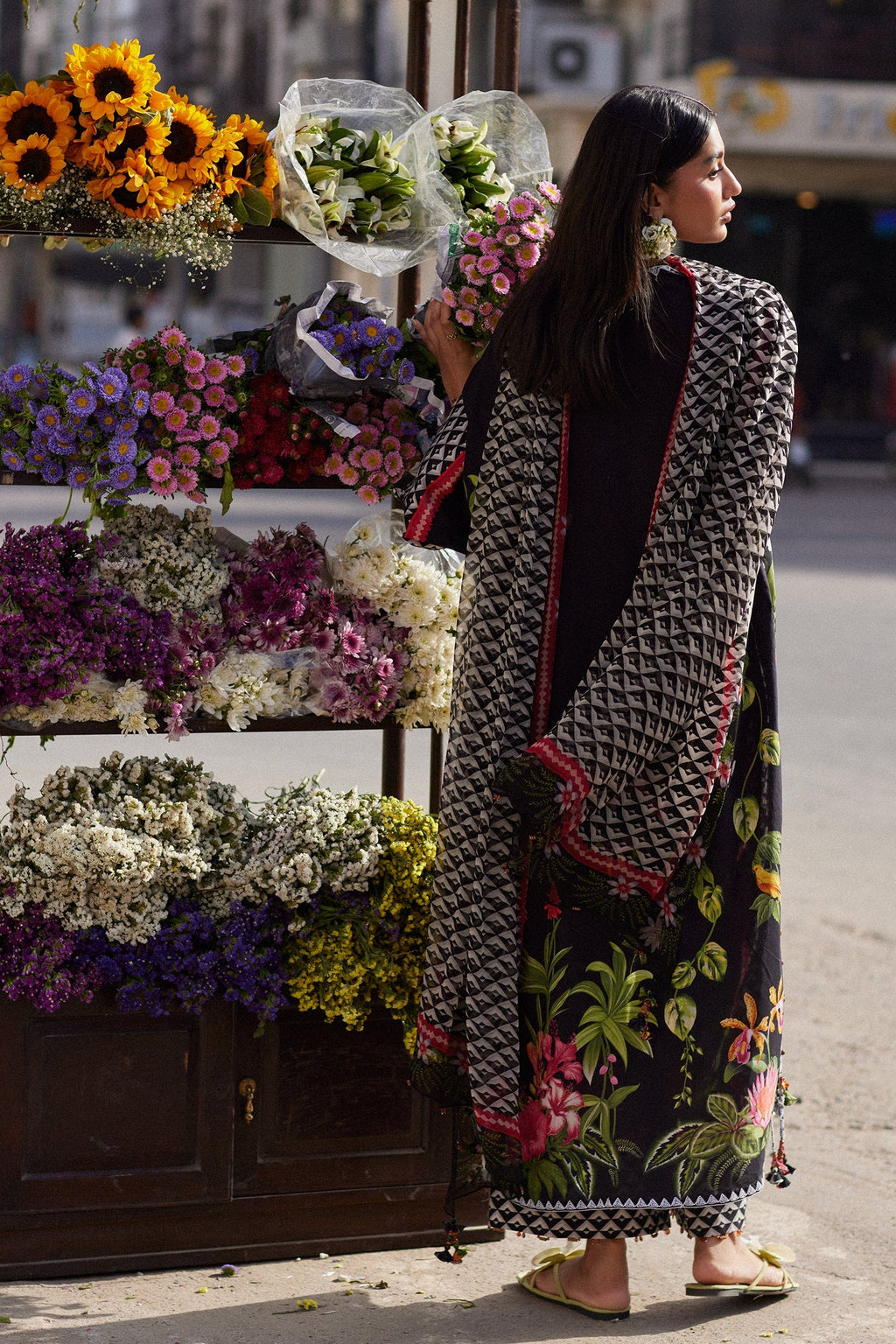 Woman in a floral dress and patterned shawl standing next to a flower stand.