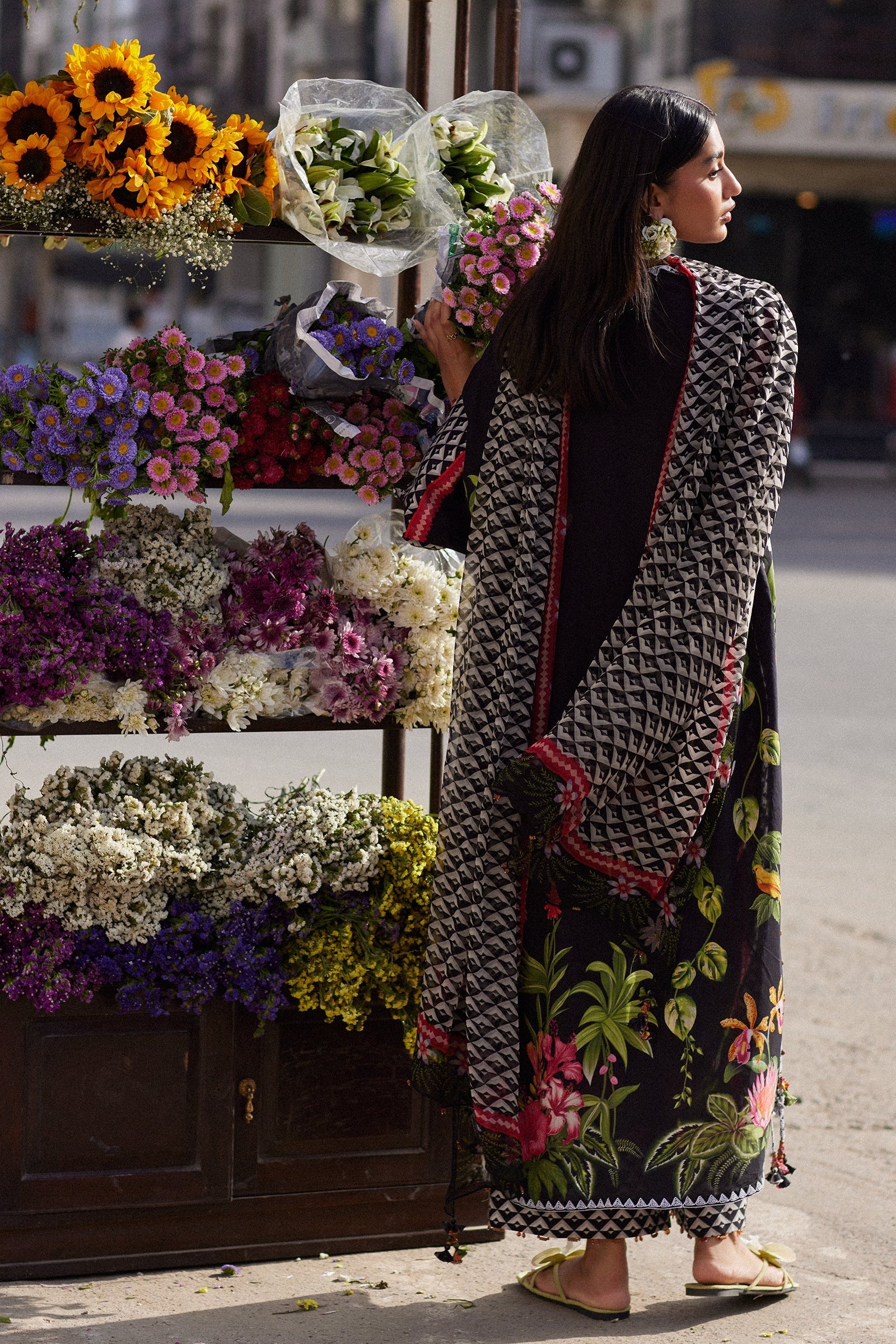 Woman in a floral dress and patterned shawl standing next to a flower stand.