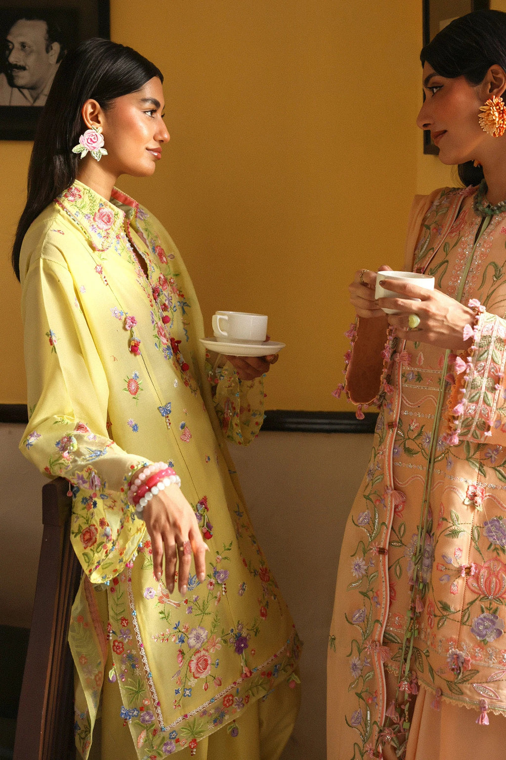 Two women in traditional outfits with floral patterns, one holding a cup, against a yellow wall.