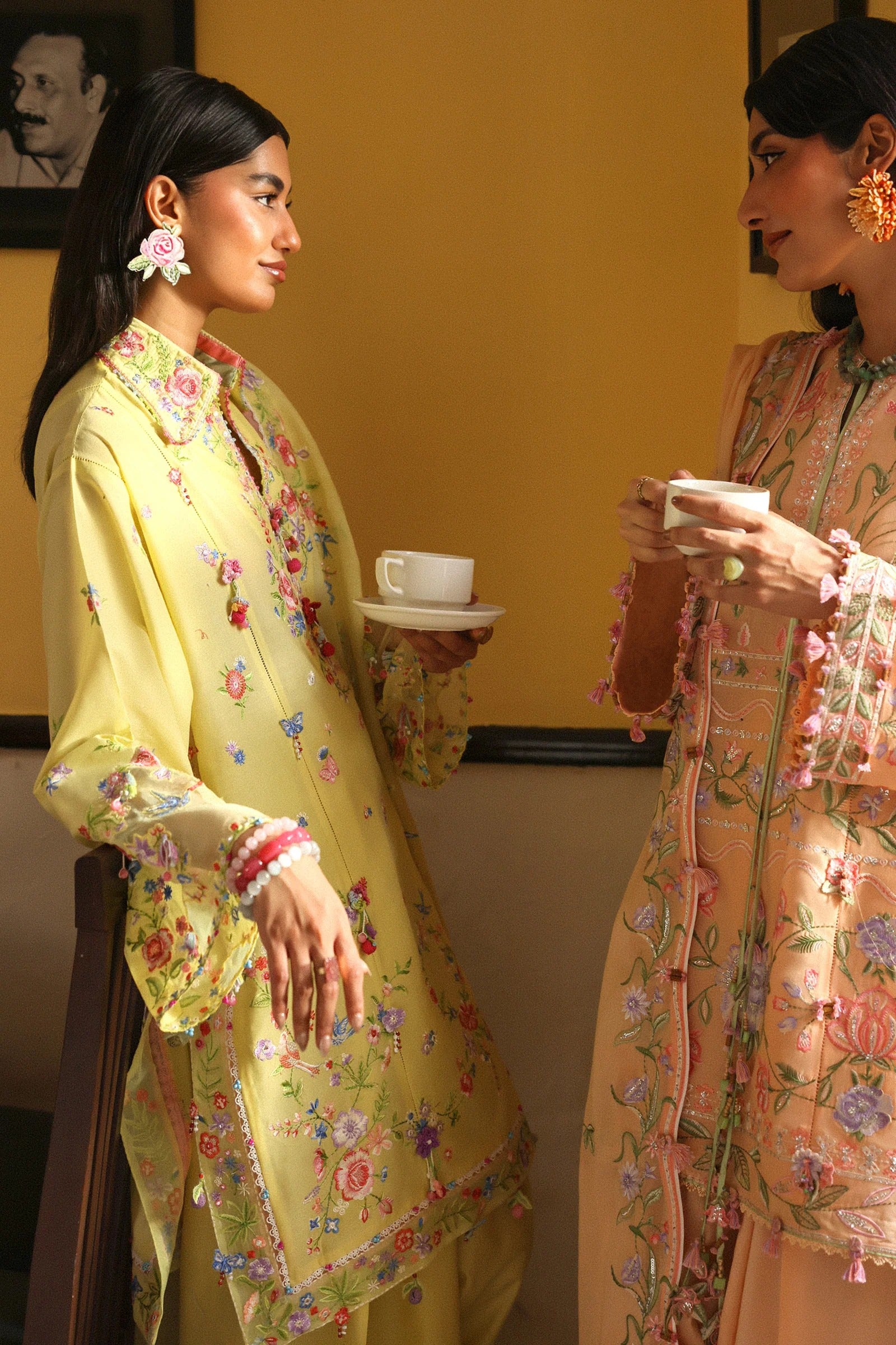 Two women in traditional outfits with floral patterns, one holding a cup, against a yellow wall.