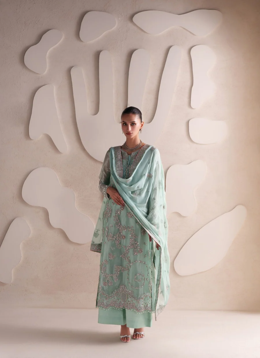 A woman stands in a studio wearing a mint green traditional outfit with floral embroidery and a matching dupatta, posed against a beige textured wall with abstract white shapes.