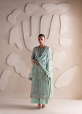 A woman stands in a studio wearing a mint green traditional outfit with floral embroidery and a matching dupatta, posed against a beige textured wall with abstract white shapes.