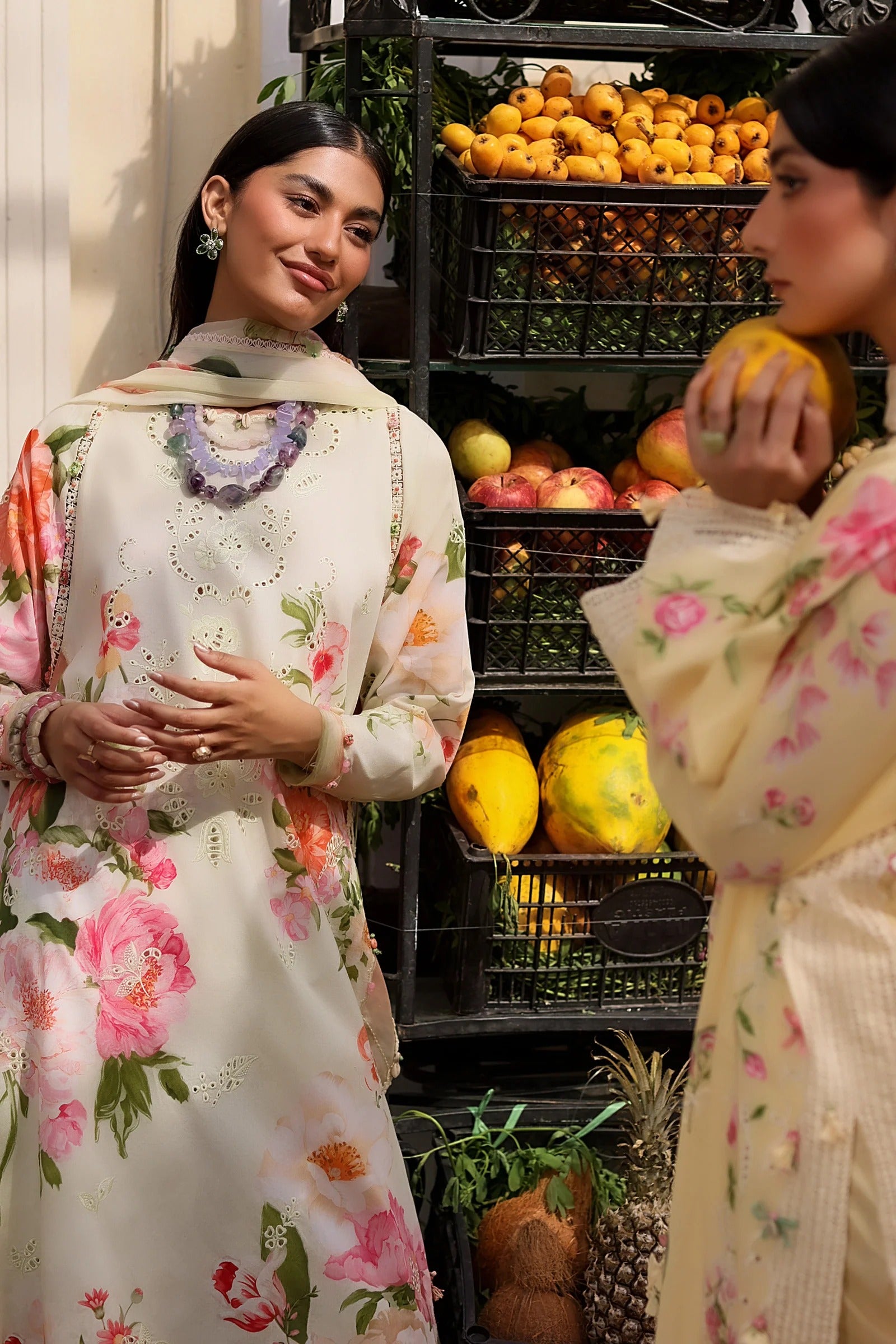 Two women in floral dresses standing in front of a fruit stand with various fruits.